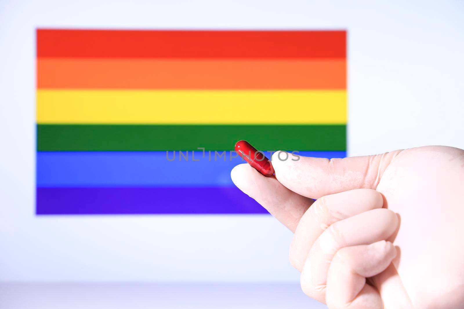 Hand in surgical glove holding red capsule and rainbow flag in the background