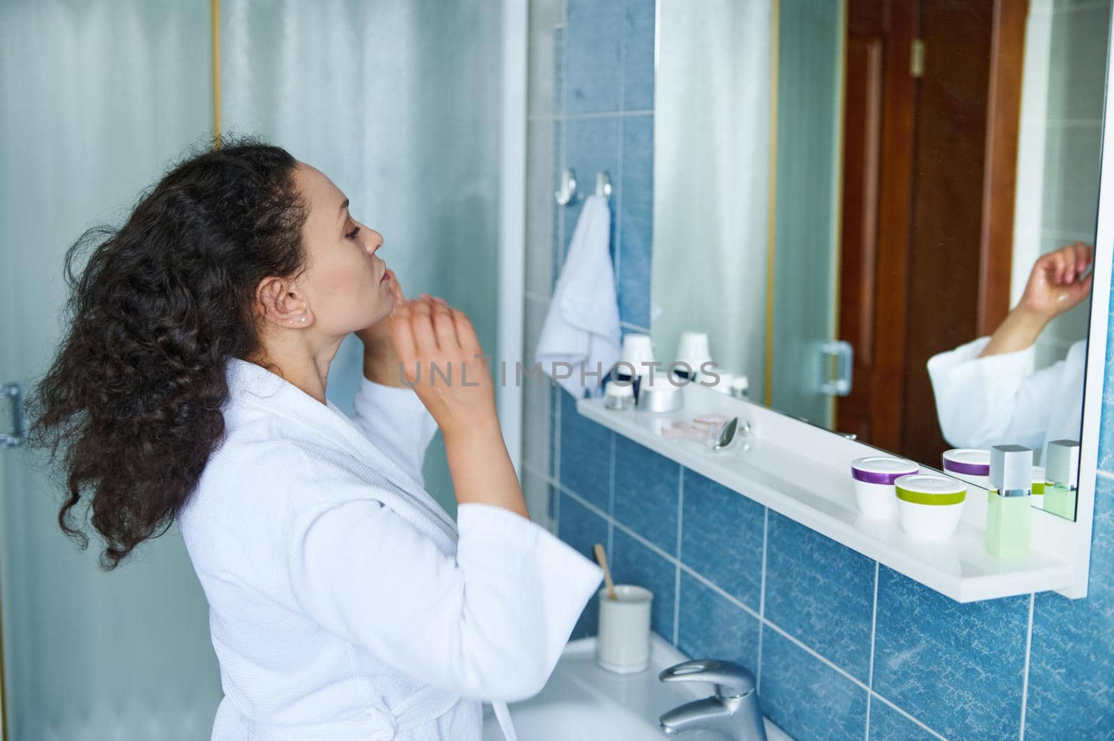 Close-up multiethnic attractive 35s woman in white bathrobe, standing in bathroom reflected in mirror, touching curly hair looking pensive thinks about hairstyle change. Beauty morning routine concept