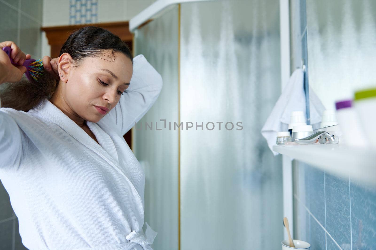 Multiethnic dark haired pretty woman brushing her curly hair with comb, standing in front of of a mirror in a home bathroom interior. Daily morning routines, hair care concept.