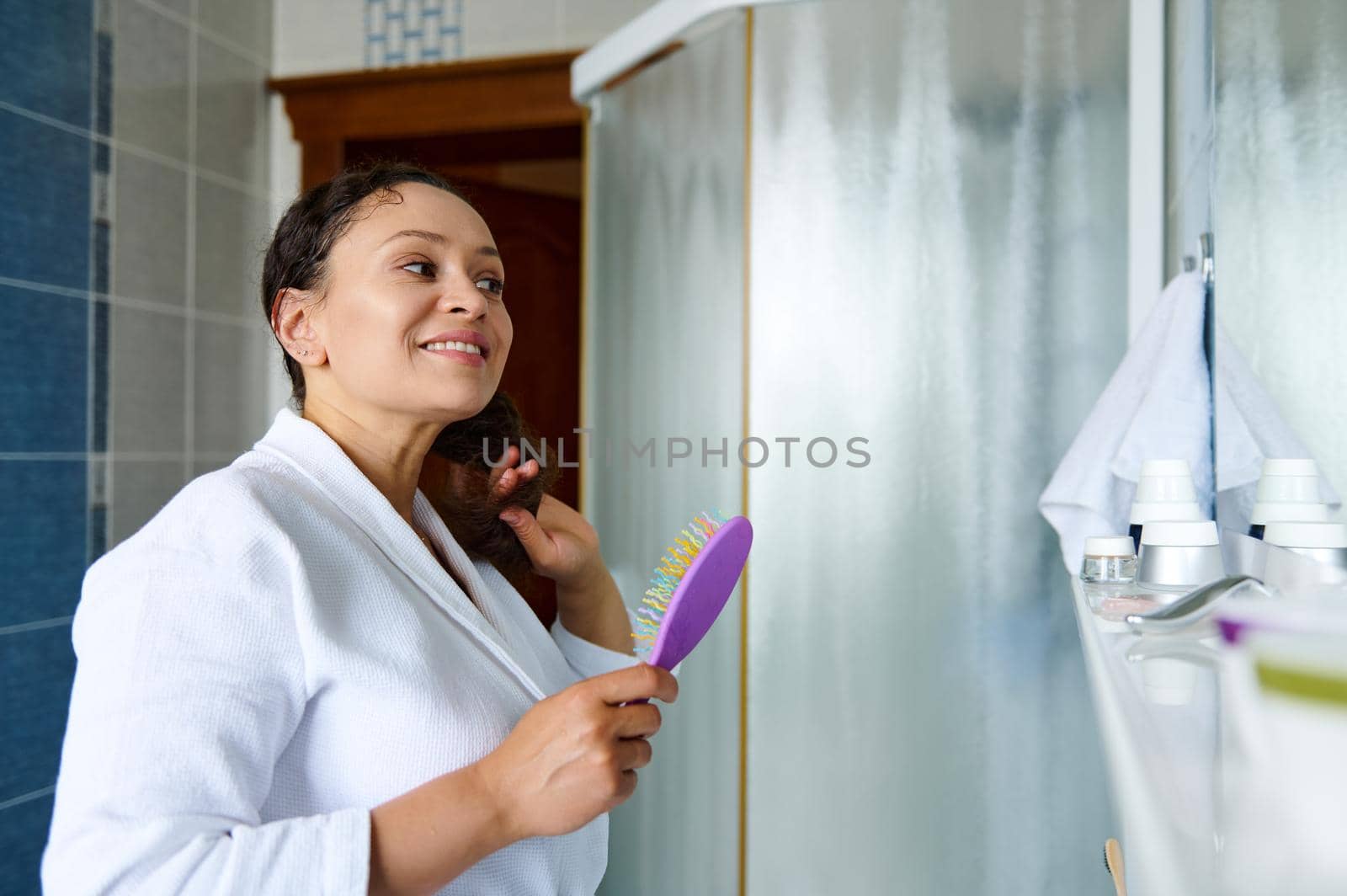 Multiethnic dark haired pretty woman brushing her curly hair with comb while looking at her reflection and smiling in mirror in a home bathroom interior. Daily morning routines, hair care concept.