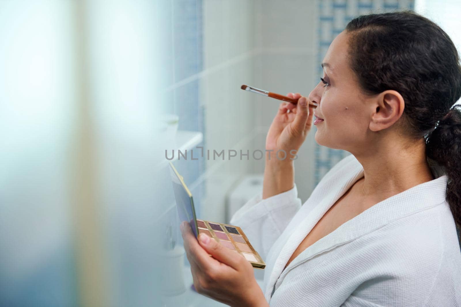 Attractive multiethnic woman, in white bathrobe, holding a colorful makeup palette, smiles looking at her reflection in the mirror, while applying eye shadows to upper eyelid, standing in the bathroom