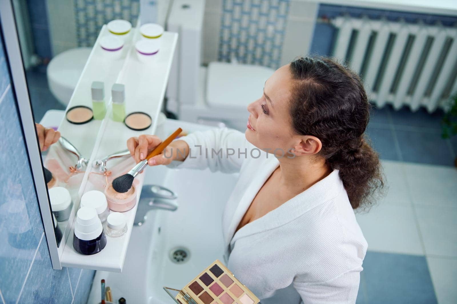 Top view of an attractive 35s woman, wearing a white bathrobe holds a colorful makeup palette and applies a mattifying powder on her face and neck, looking at her reflection in the bathroom mirror