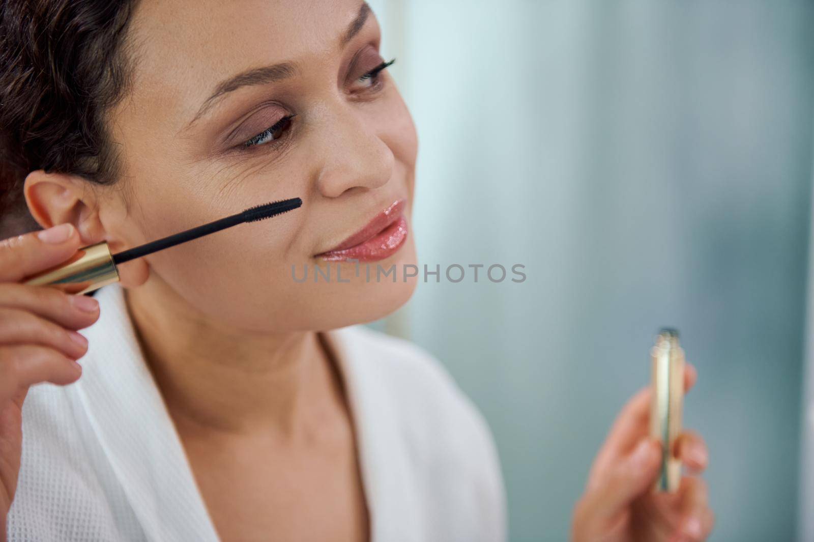 Headshot of a stunning young 30s multiethnic woman in white bathrobe, with perfect make-up adding volume to her eyelashes, applying mascara, while standing in front of the mirror in the home bathroom