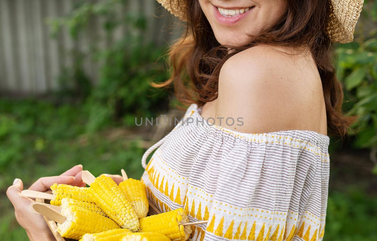 The girl is smiling in a straw hat and holding a plate with boiled corn in her hands. The concept of outdoor recreation, barbecue