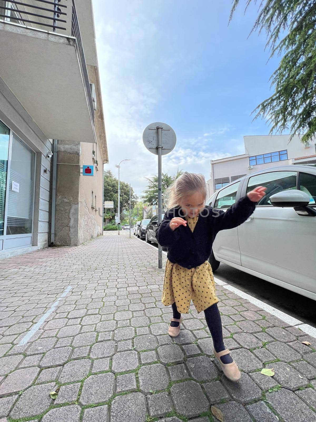 Little girl walks along the paving stones past a parked car by Nadtochiy