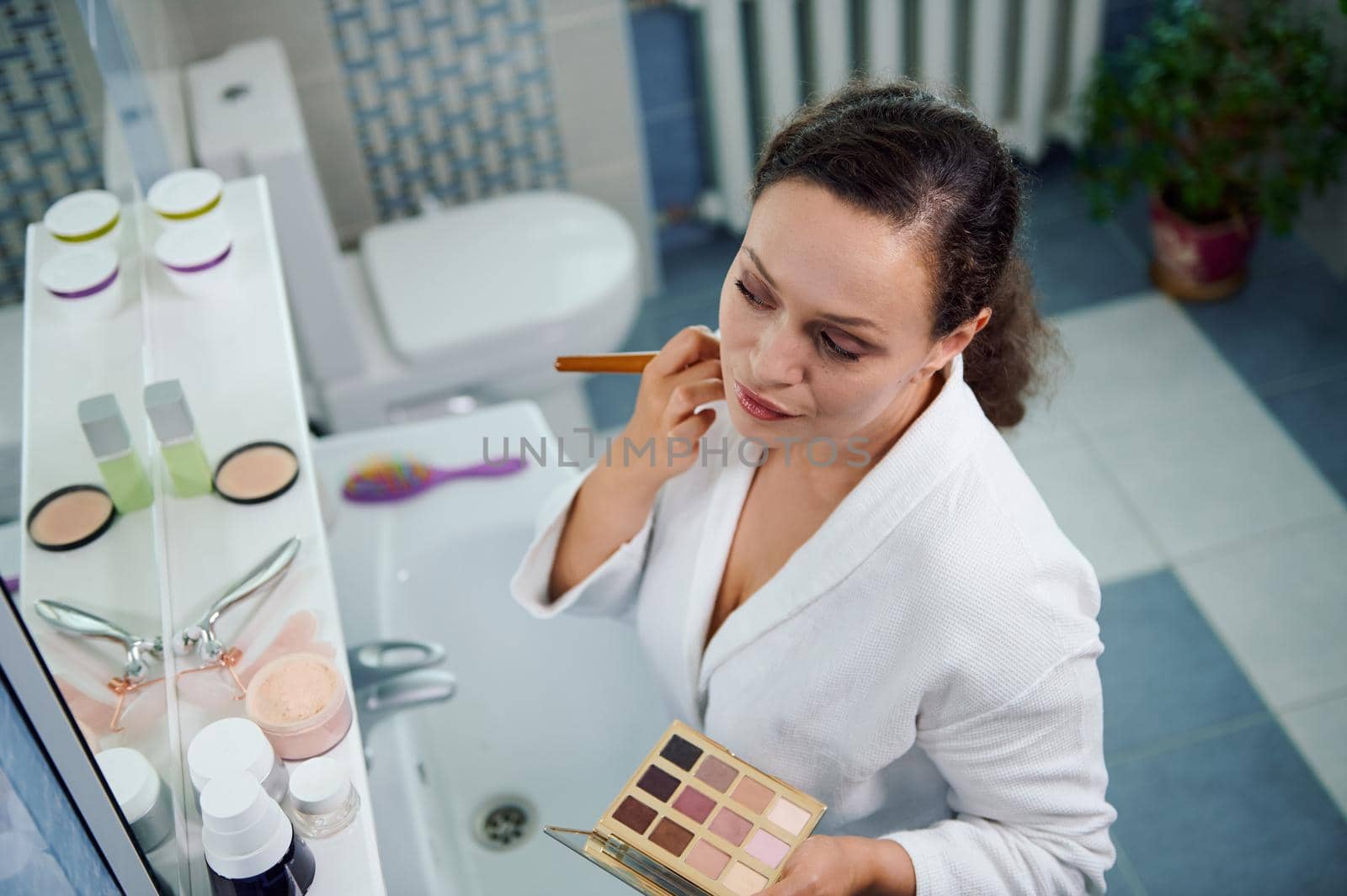 Pretty, stunning woman, in white bathrobe holds eyeshadow palette, applies mattifying powder on her face, satisfied of her appearance and natural make-up looking at her reflection in bathroom mirror