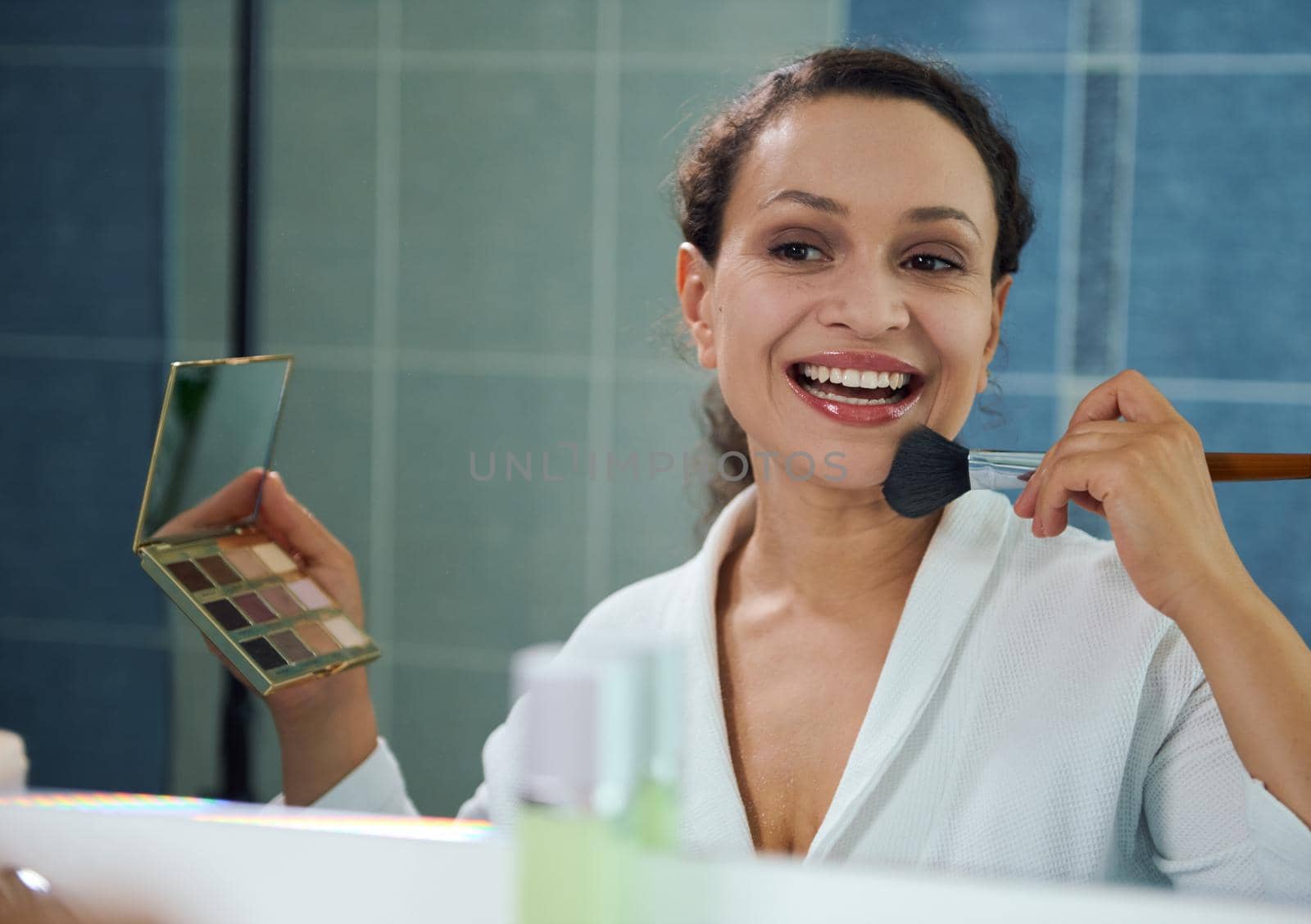 Attractive young woman, reflecting in mirror, holds eyeshadow palette, smiles a beautiful toothy smile while applying a mattifying powder on her face, satisfied of her appearance and natural make-up