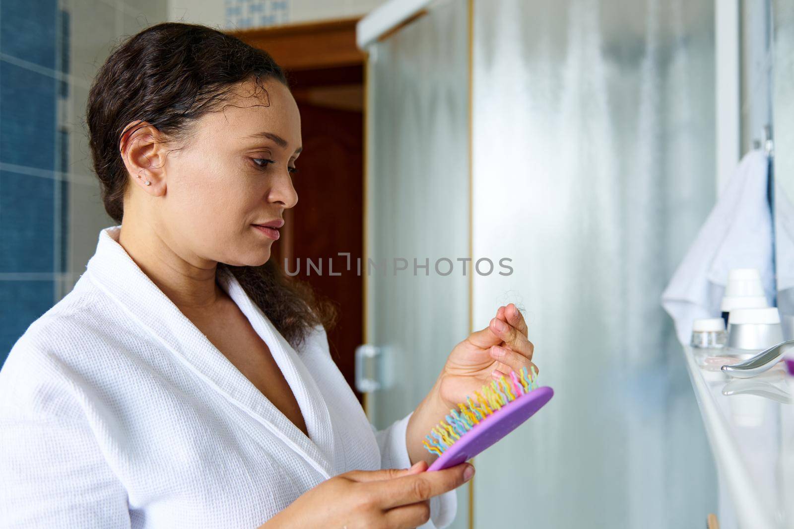 Multiethnic dark-haired woman looks in the mirror while combing her curly hair. She is irritated and dissatisfied with the state of damaged dry hair. The problem of curly hair, split ends, hair loss.