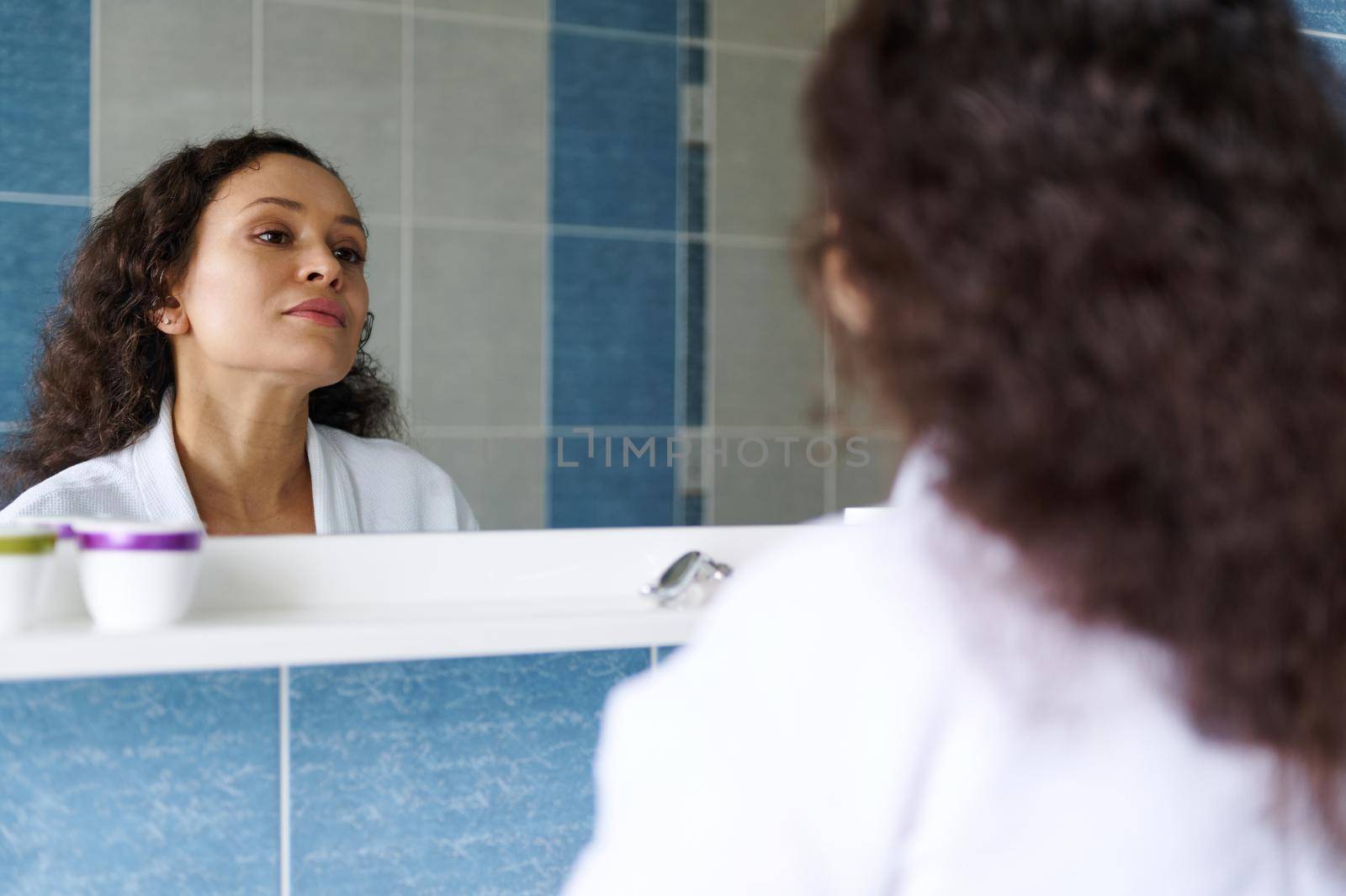 Middle-aged beautiful multi-ethnic woman in a white waffle bathrobe, admires herself in the mirror while performing morning beauty treatments in the bathroom. Skin and body care concept. Aging process