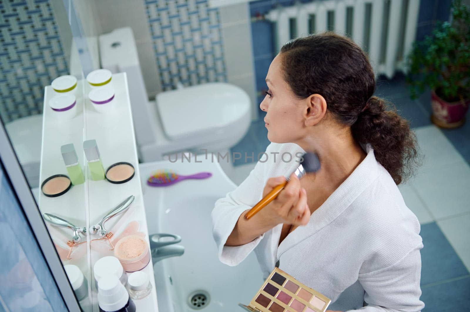 Top view of a stunning multiethnic woman, wearing a white bathrobe holds a colorful makeup palette and applies mattifying powder on her face and neck, looking at her reflection in the bathroom mirror