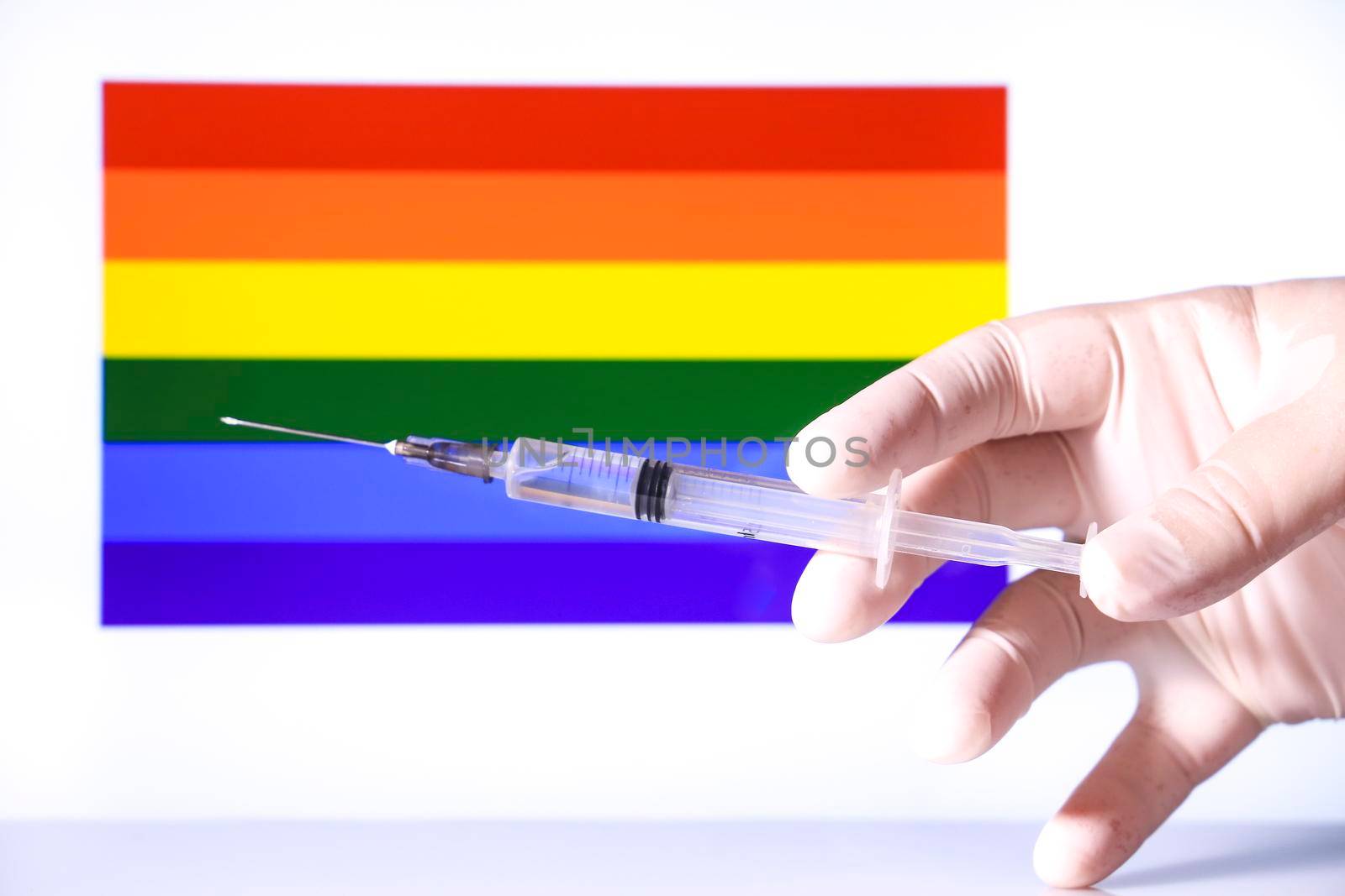 Hand in surgical glove holding syringe and rainbow flag in the background