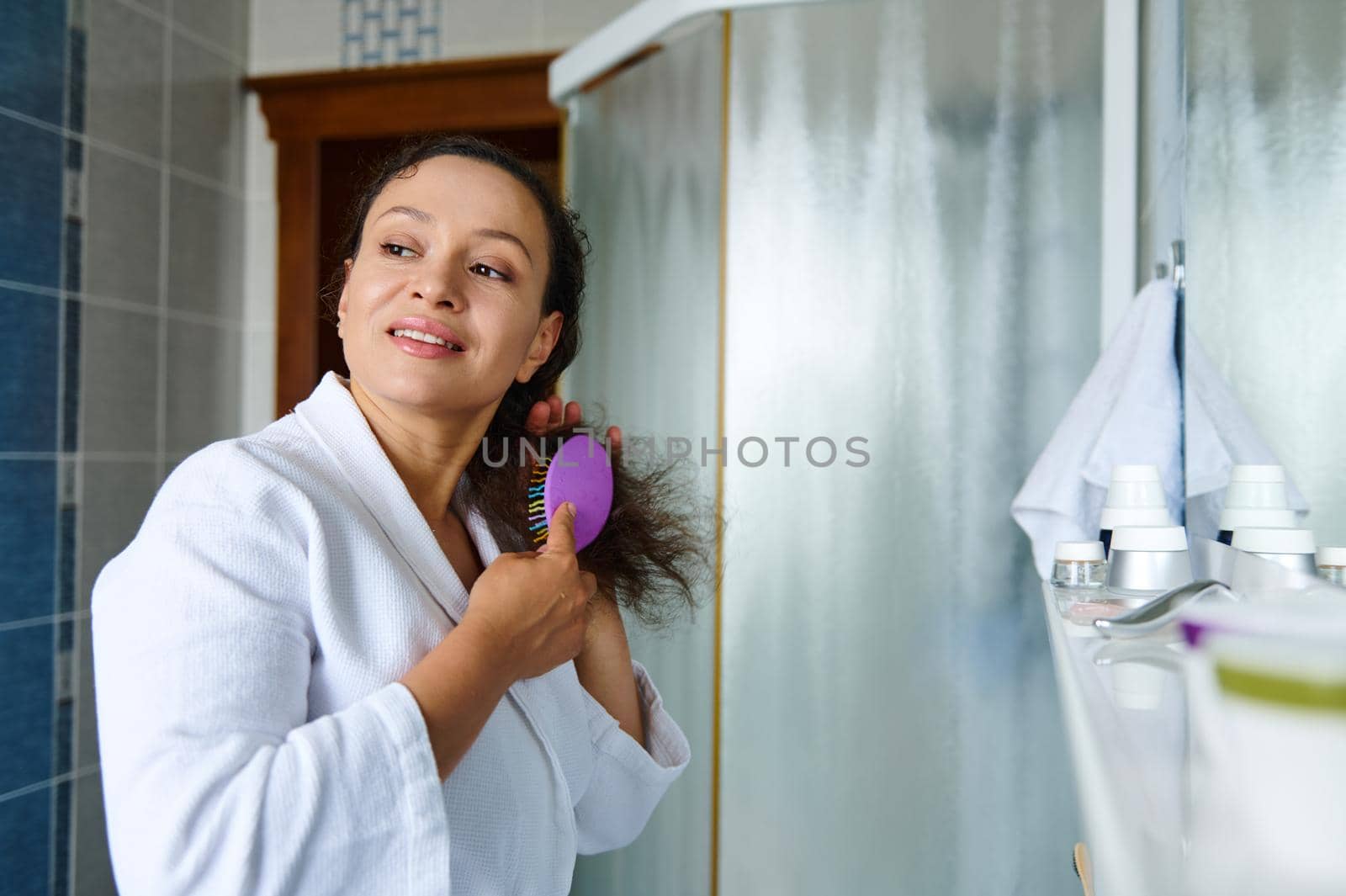 Beautiful multiethnic woman, in a white waffle bathrobe, combing hair, looking aside while caring for curly chevelure, during morning routines, in the bathroom. Beauty and body care concept