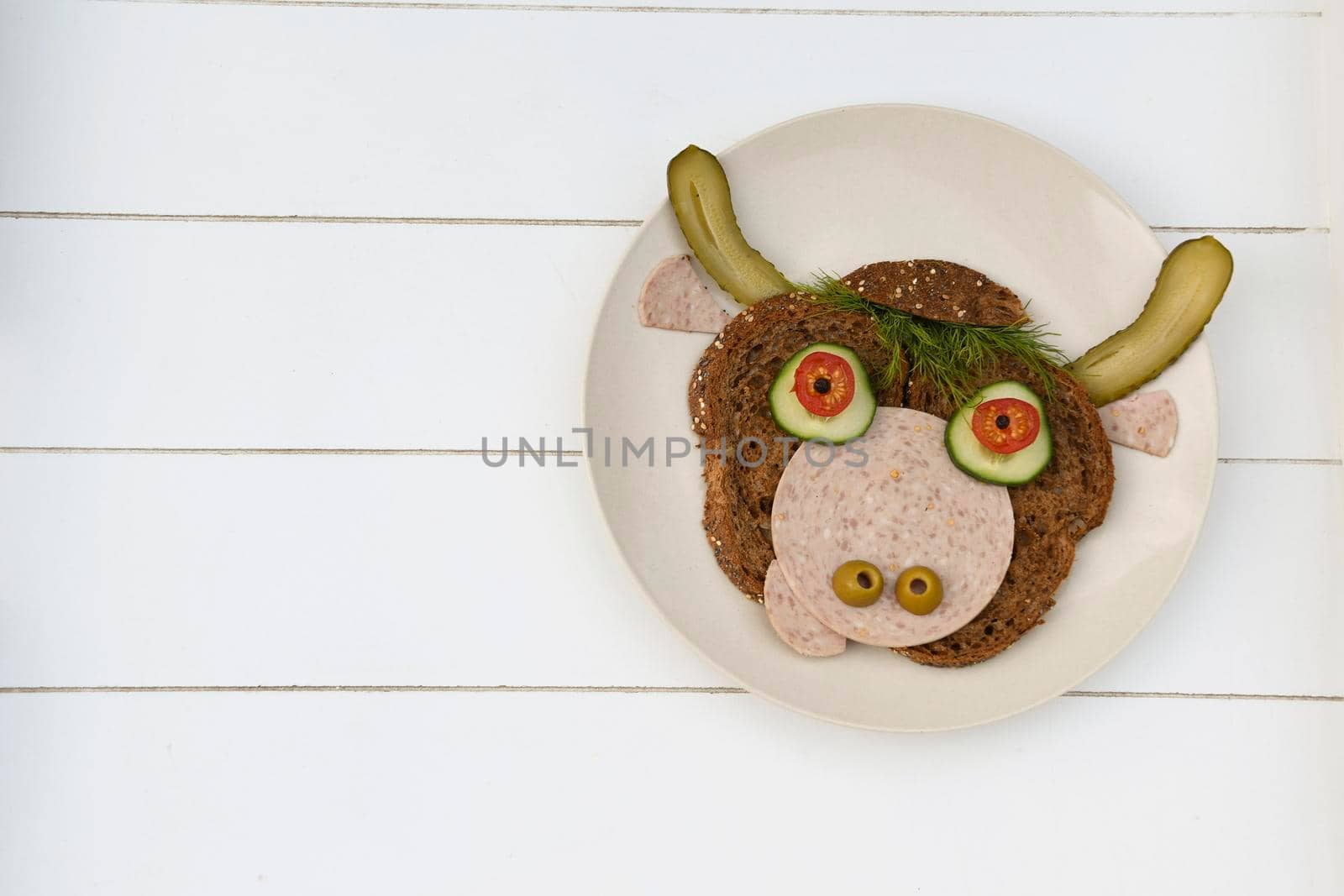 Sandwich in the form of a portrait of a cheerful bull made of dark bread, sausages and vegetables on a plate, on a white wooden background, copispice. High quality photo