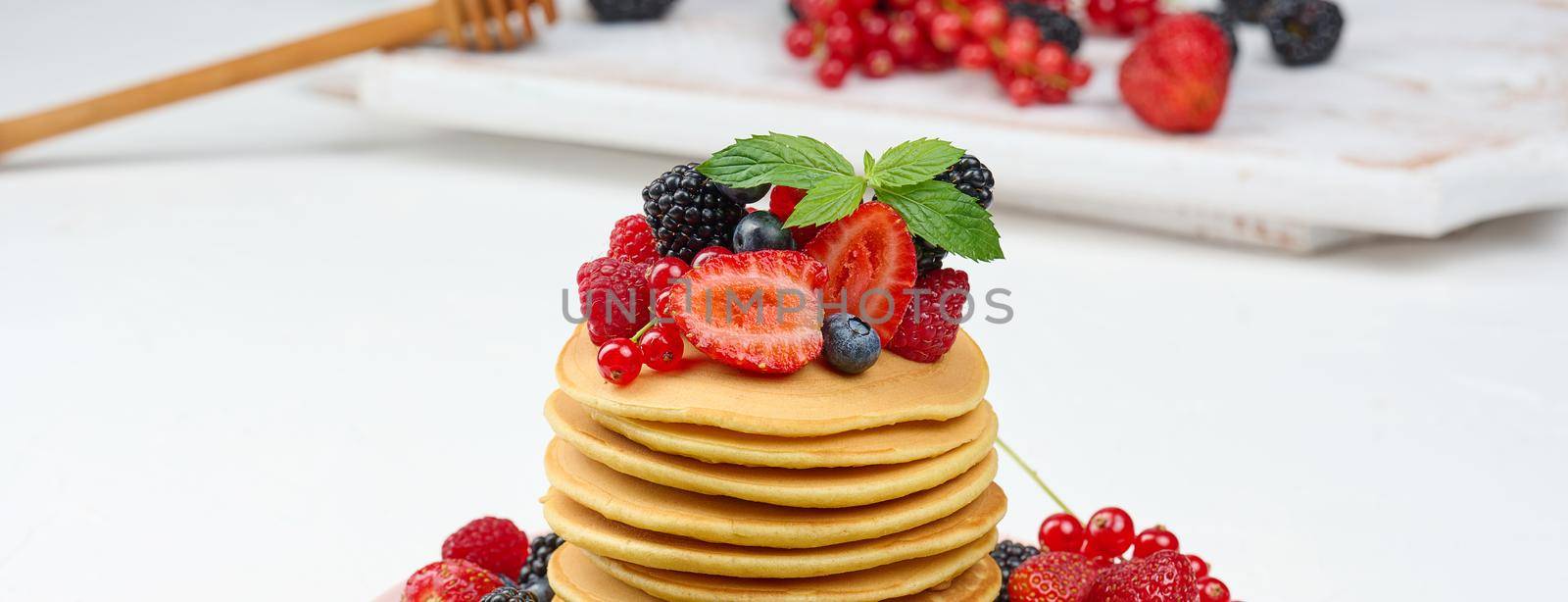 Stack of baked pancakes with fruits in a round plate on a white table, banner
