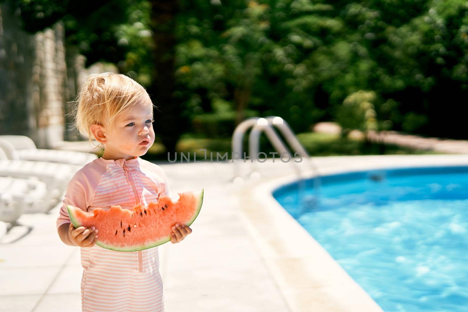 Little girl with a piece of watermelon in her hands stands near the pool with turquoise water. High quality photo