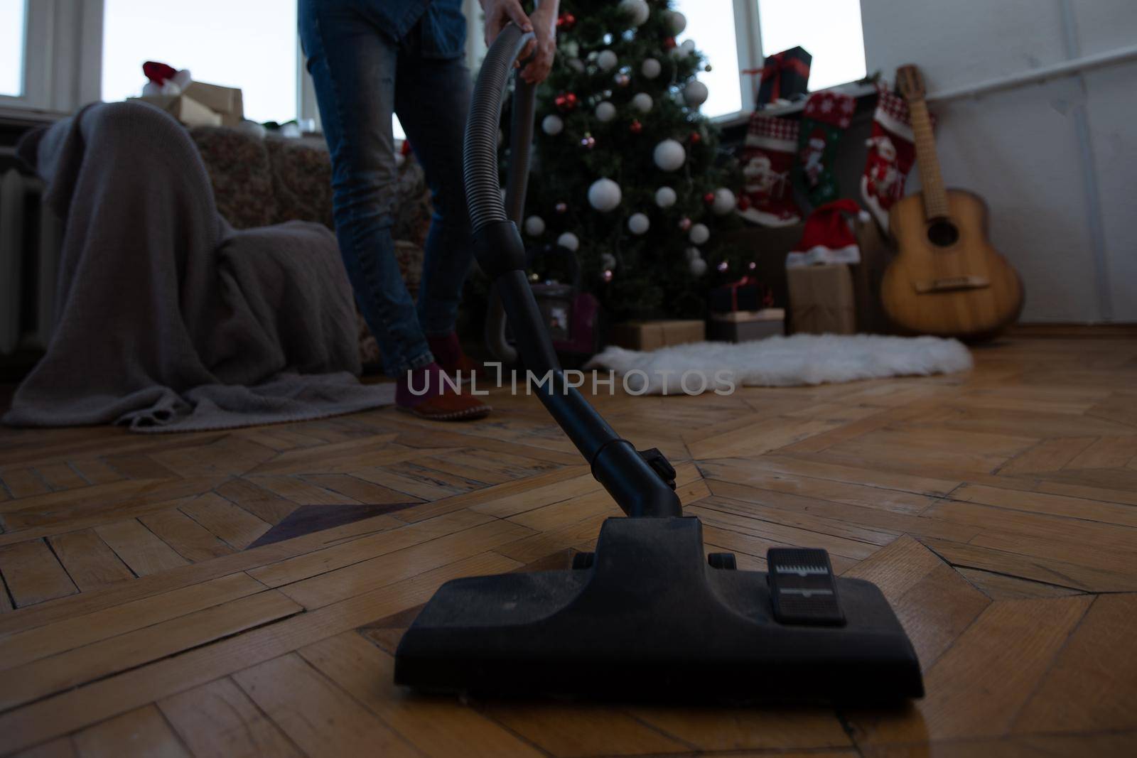 Young woman cleaning with vacuum cleaner, vacuuming under Christmas Tree needles with New Years ornaments on hardwood wooden floor