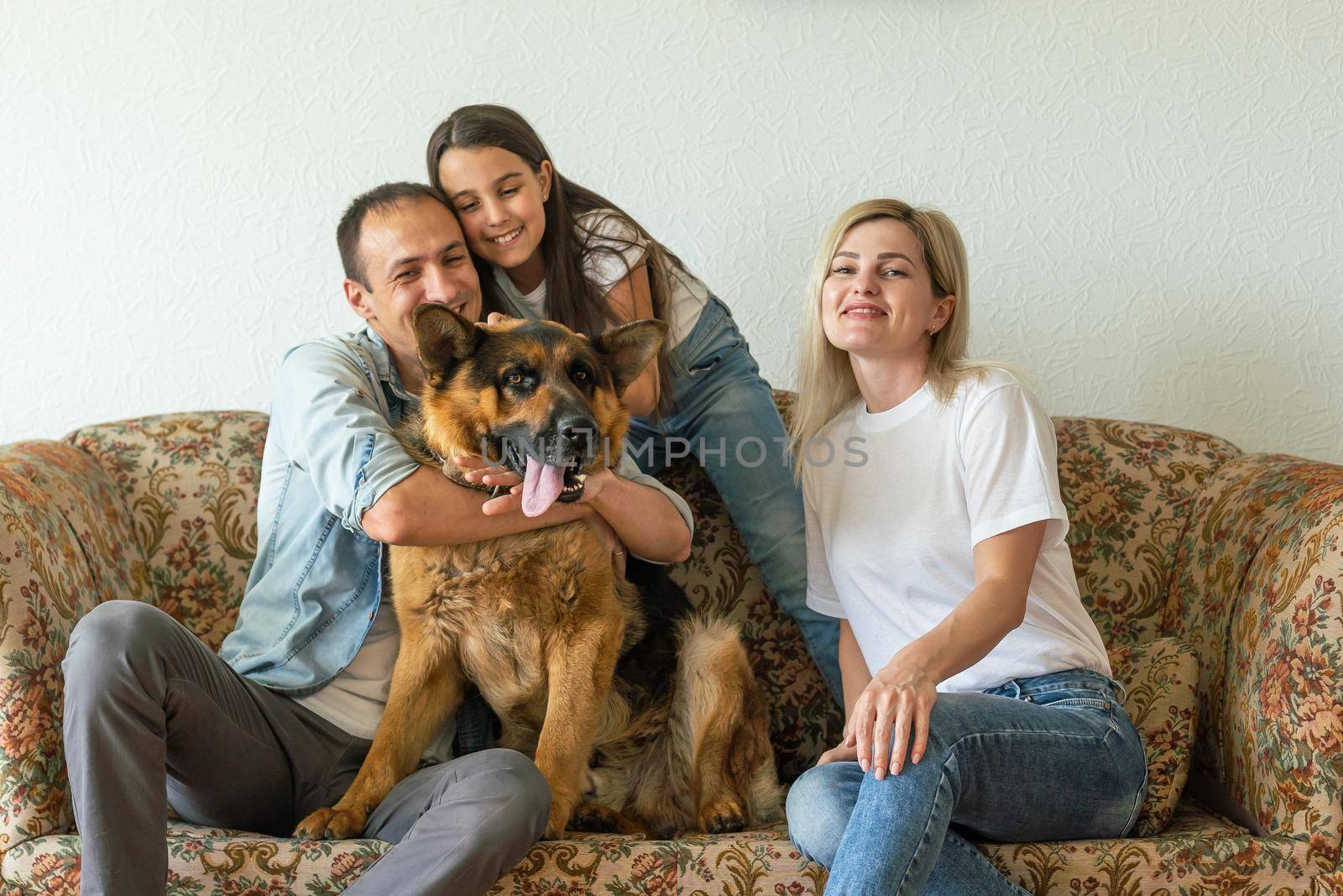 Beautiful young parents, their cute little daughter looking at camera and smiling, sitting with their cute dog on sofa at home.