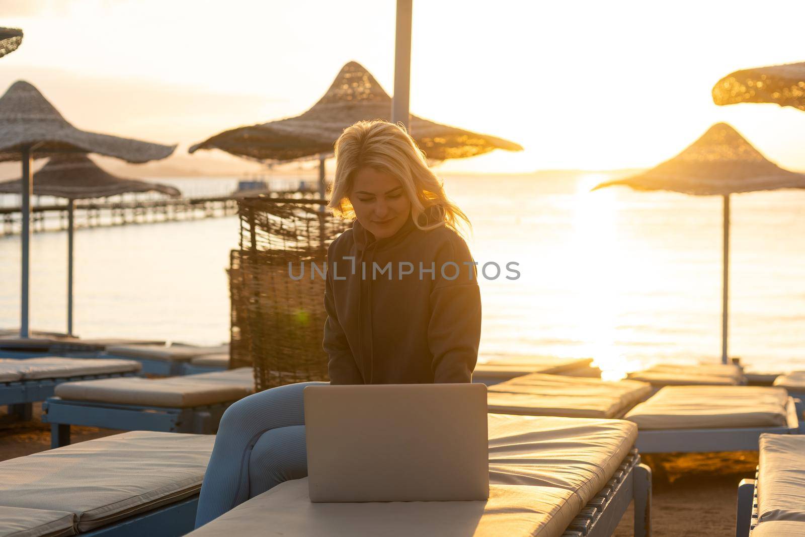 Working remotely on seashore. Young successful woman female freelancer in straw hat working on laptop while sitting on tropical beach at sunset, full length. Distance work concept. by Andelov13