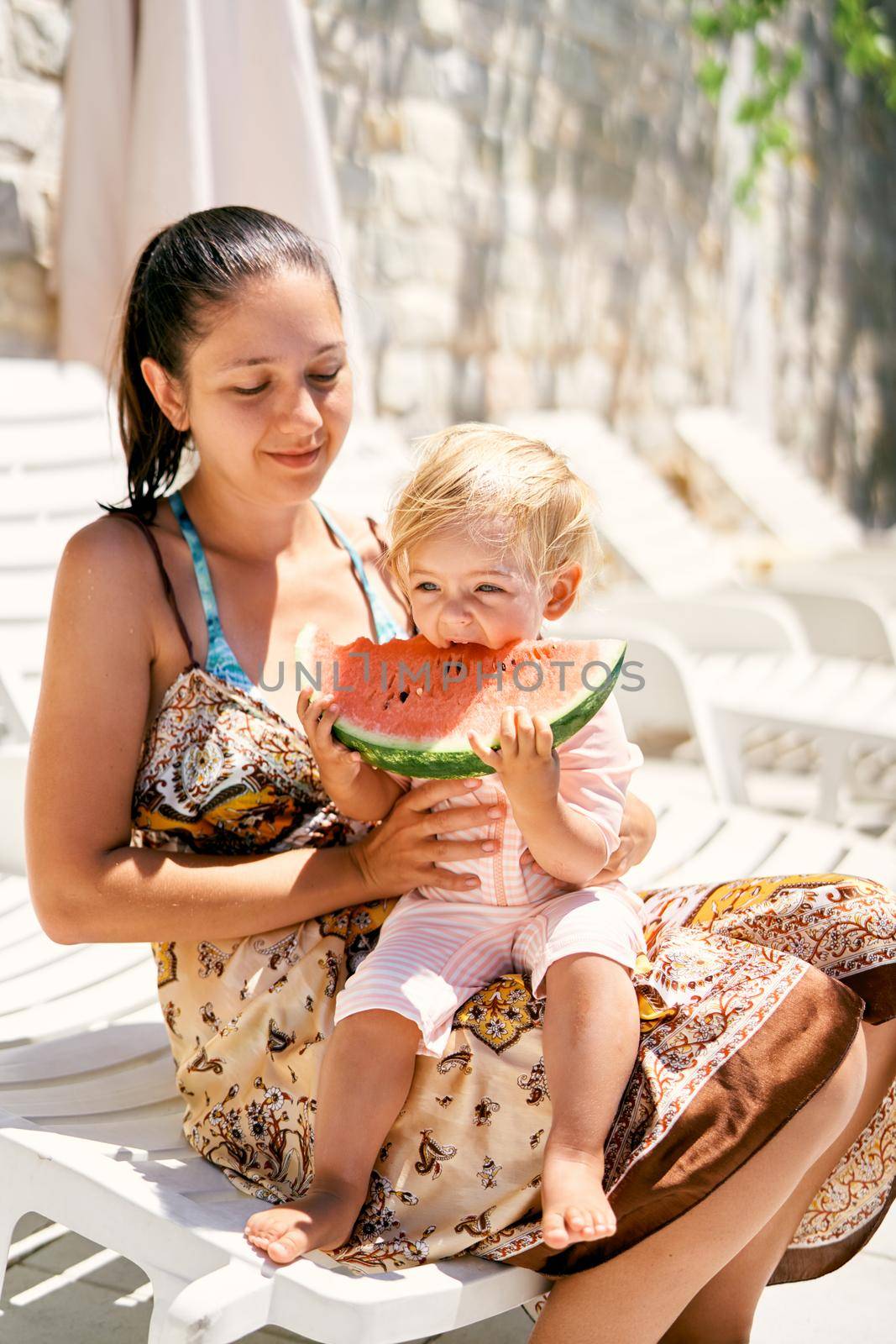 Little girl eats a piece of watermelon while sitting in her mother arms by Nadtochiy