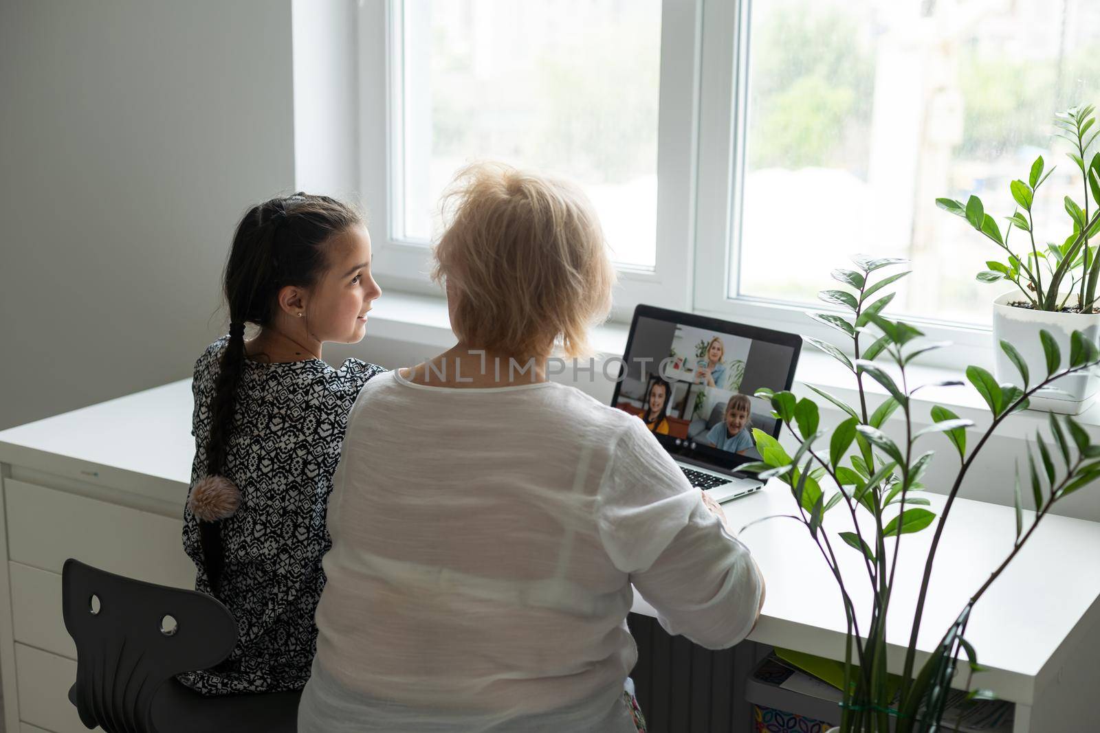 Portrait of happy grandmother and little granddaughter making video conference on pc sitting at table, waving hands at screen, greeting somebody, chatting with parents, enjoying online communication. by Andelov13