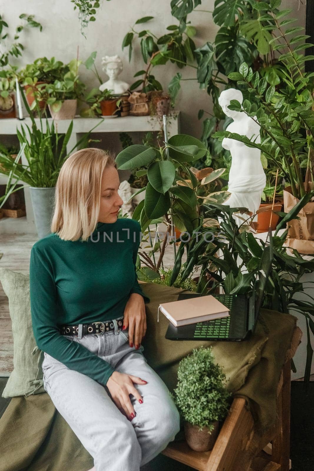 A blonde woman in a room with a lot of green indoor plants is working on a laptop. The concept of biophysical design in the interior. Work from home, work as a freelancer.