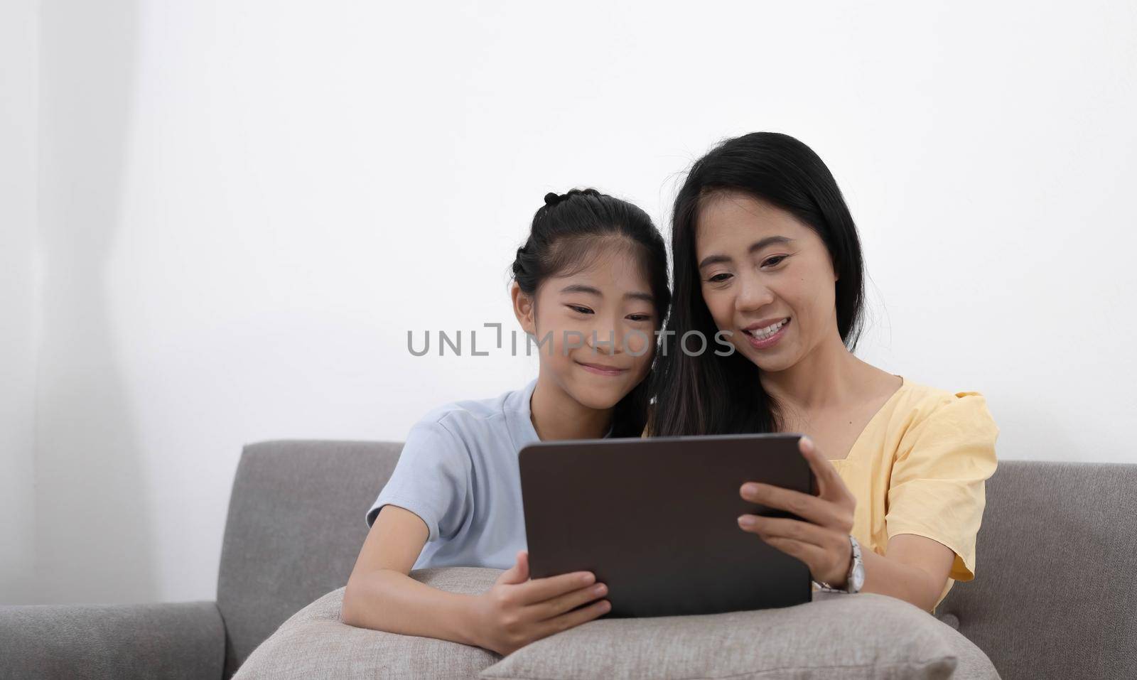 Happy young asian mom and daughter using digital tablet, watching videos or surfing internet, sitting on sofa at home, copy space. by wichayada