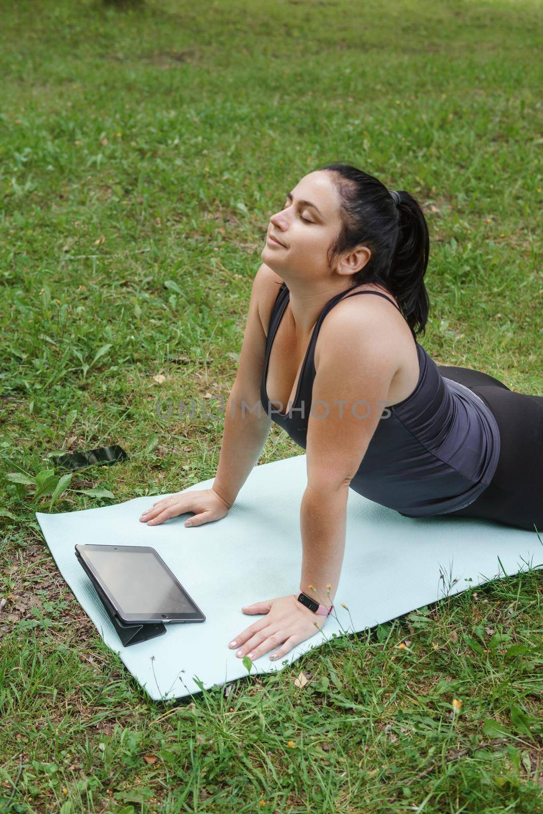 A charming brunette woman plus-size body positive practices sports in nature. Woman does yoga in the park on a sports mat. On a yoga mat in the dog pose.