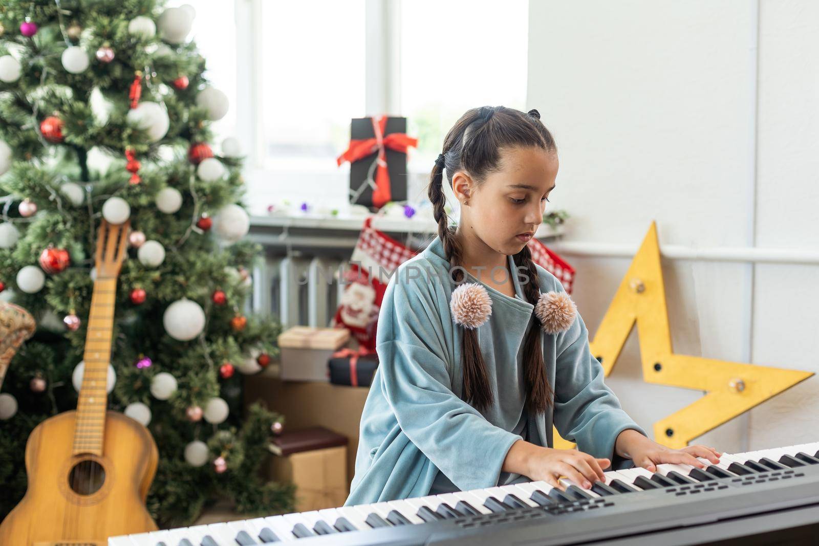 A cute little girl sitting near a white piano and a Christmas tree. The concept of the New Year, family holidays