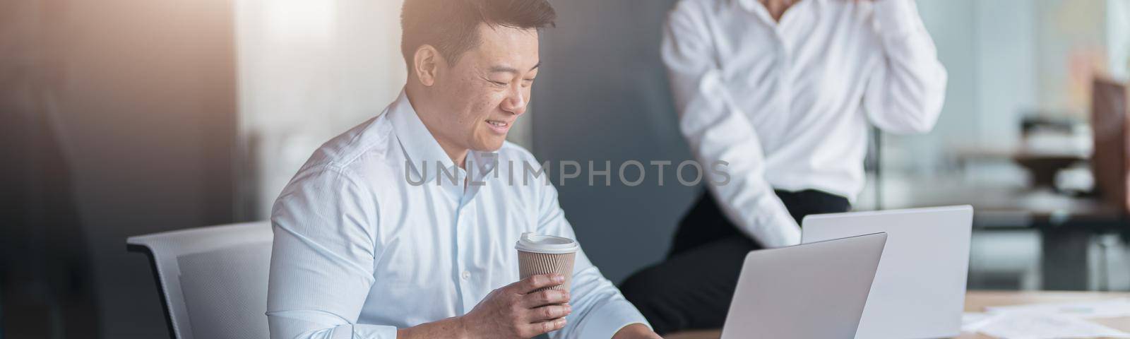 Asian businessman drink coffee and working on laptop with colleague talking on phone in background