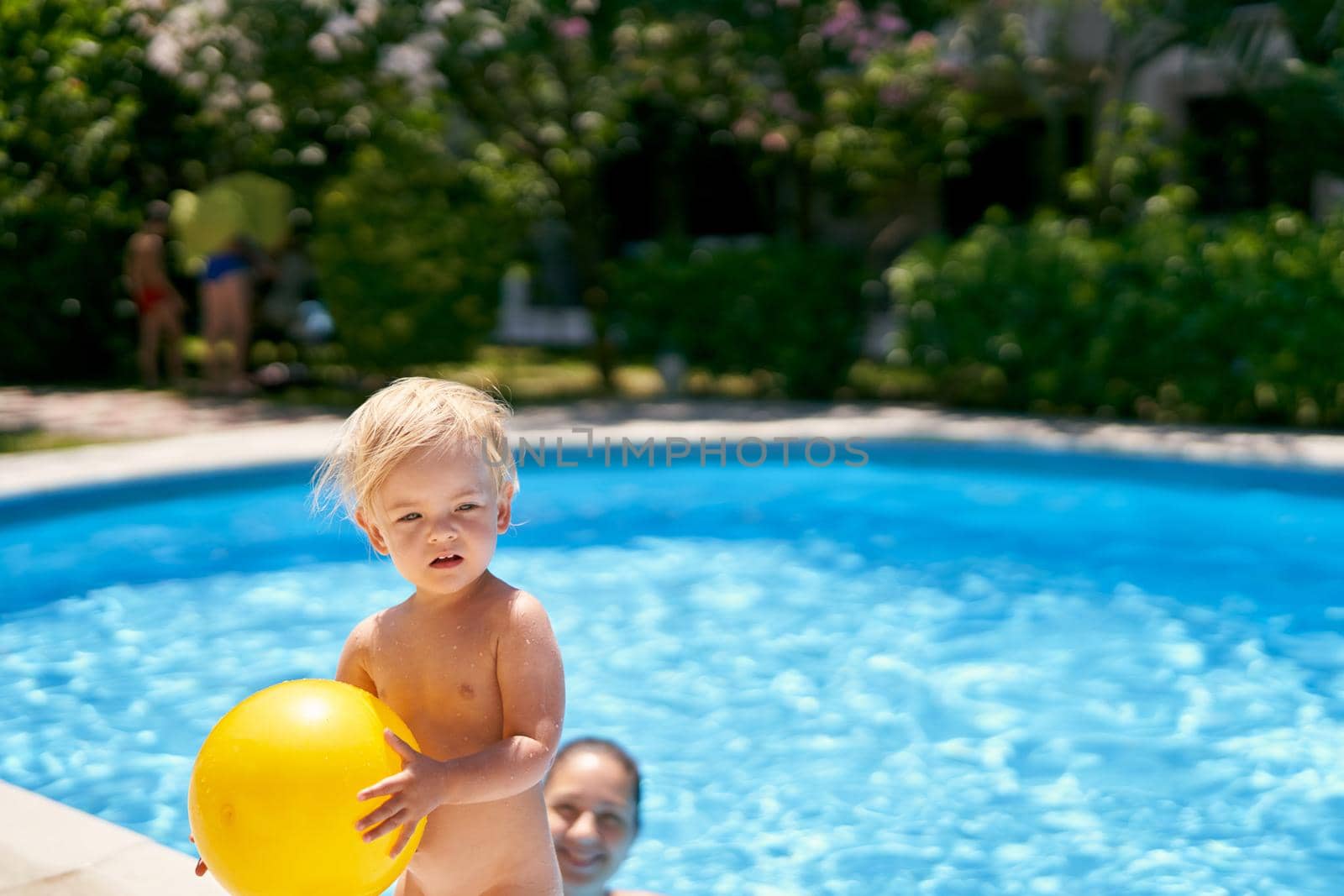 Small baby with a yellow ball stands by the pool with turquoise water. High quality photo