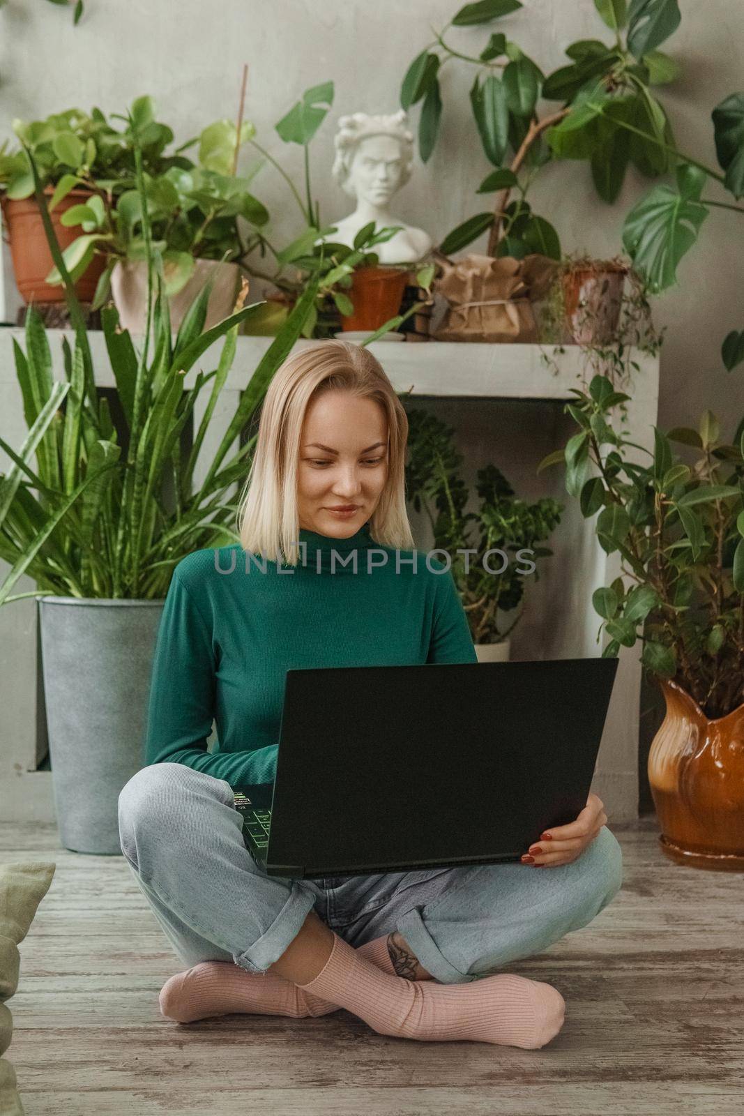 A blonde woman in a room with a lot of green indoor plants is working on a laptop. The concept of biophysical design in the interior. Work from home, work as a freelancer. by Annu1tochka
