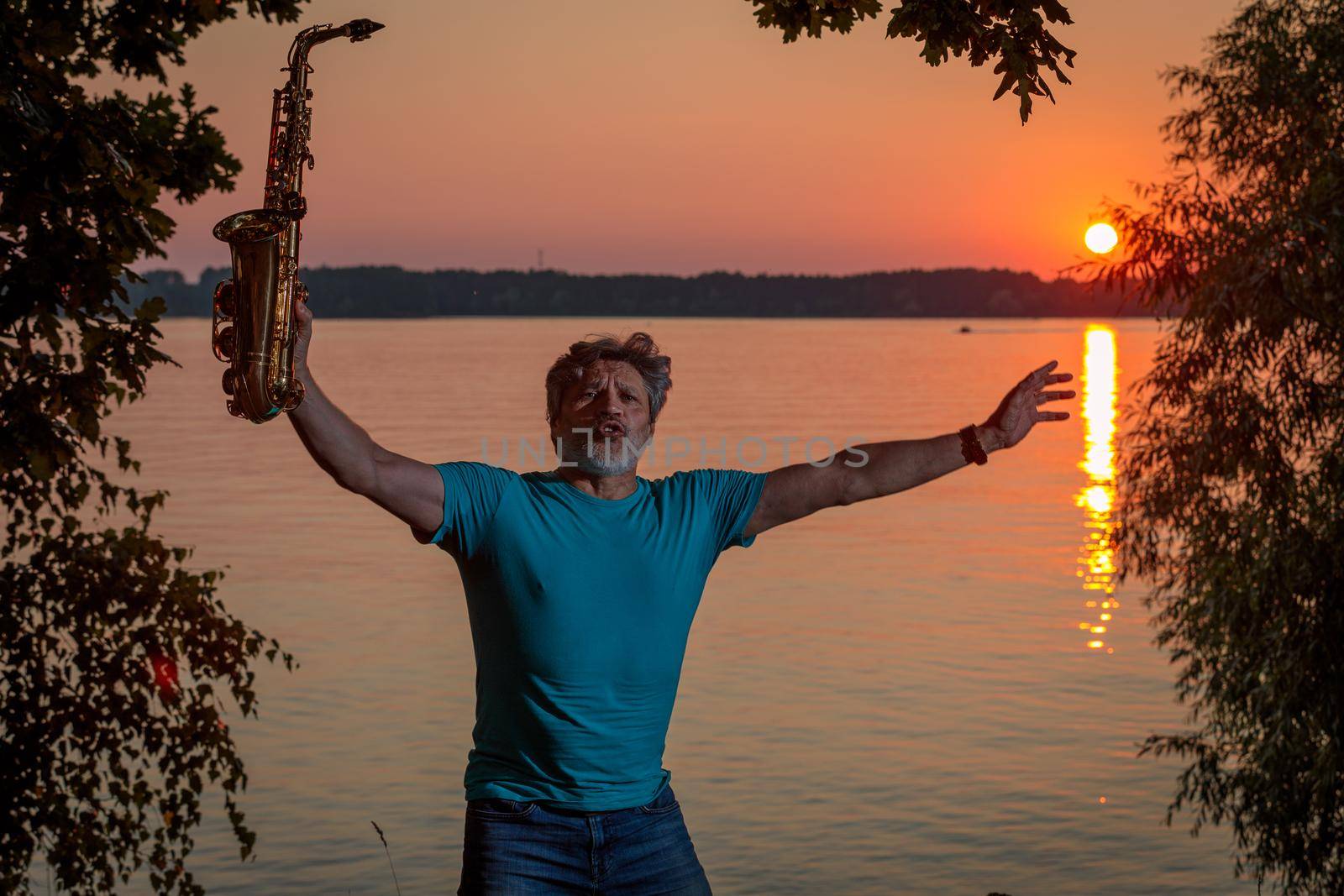 An adult man plays the saxophone at sunset by the river in the evening by Yurich32