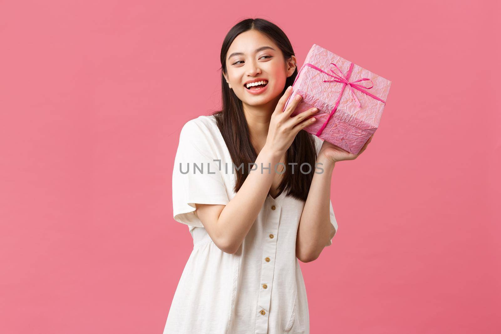Celebration, party holidays and fun concept. Curious cute excited woman in white dress celebrating birthday, wonder whats inside b-day gift, shaking box and smiling happy, pink background.