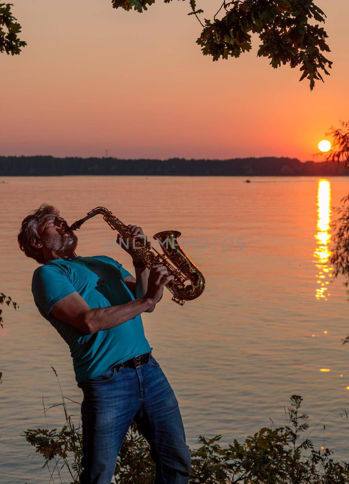 An adult man plays the saxophone at sunset by the river in the evening by Yurich32