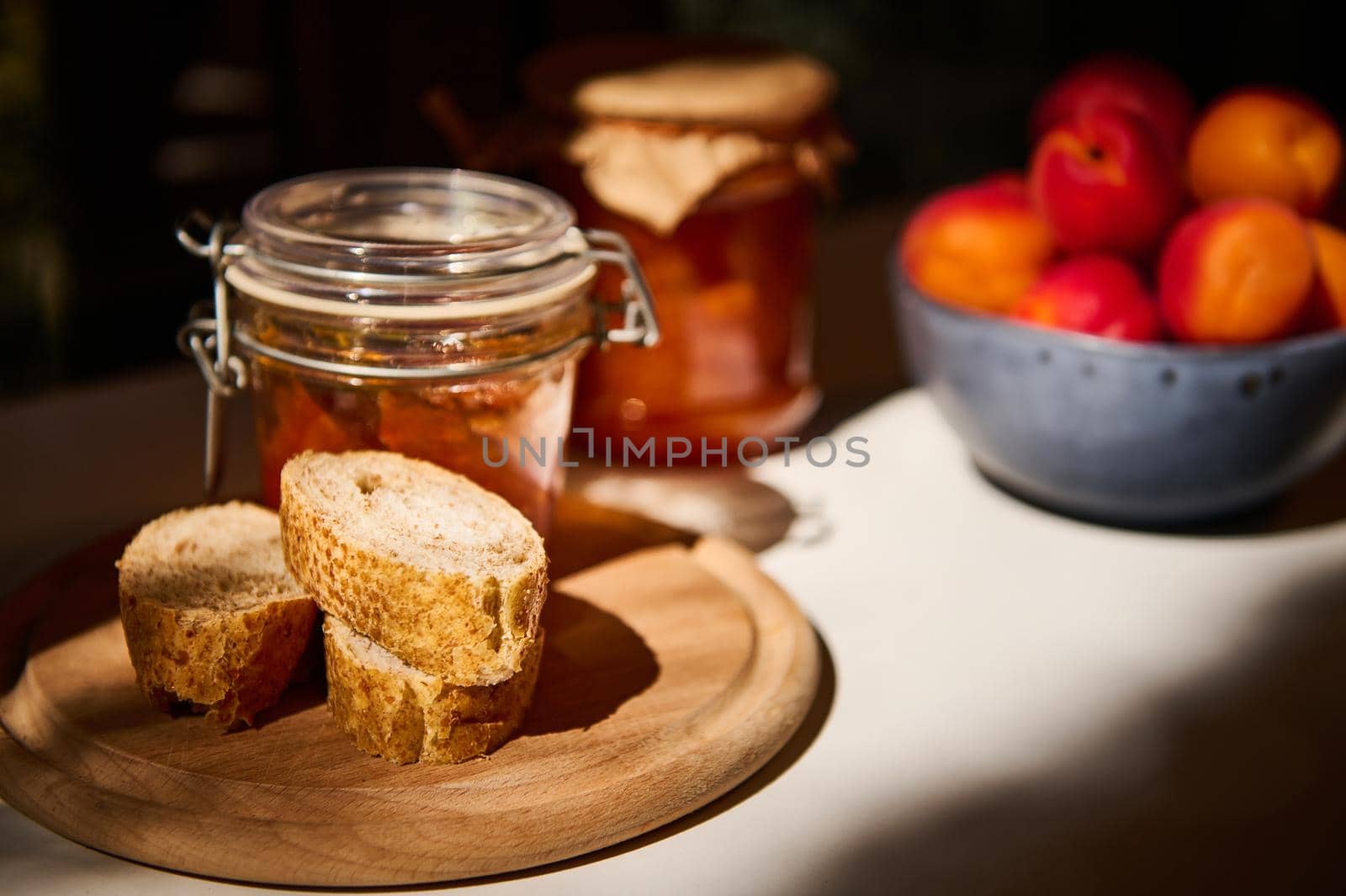 Still life, illuminated by a daylight. Close-up slices of whole grain bread on wooden board, jar with homemade jam and ripe juicy apricots in blue ceramic bowl on white table in rustic summer kitchen