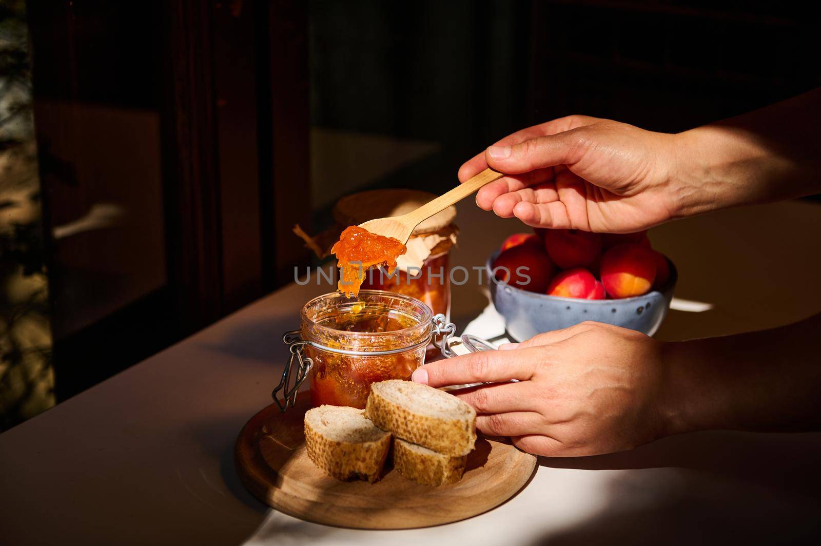 Food and drink, summer autumn harvest concept. Close-up of the hands of a housewife holding a wooden spoon and spreading homemade apricot jam on bread. Image illuminated by daylight.
