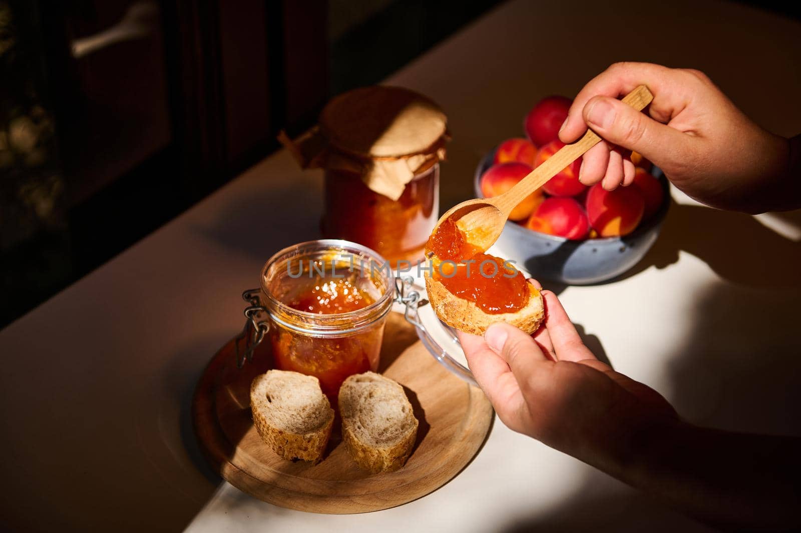 Daylight falls on the table. Hands using wooden spoon, spreads a homemade confiture on the bread. Rustic still life composition with fresh ripe apricots in a blue bowl and a jar with marmalade and jam