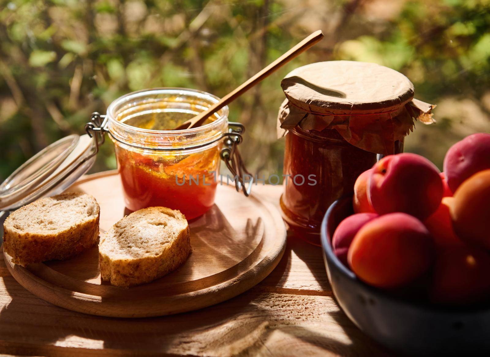 Close-up of delicious ripe red apricots in a blue ceramic bowl on a wooden table with a glass jar of homemade apricot confiture and slices of freshly baked whole grain bread. Rustic style. Still life