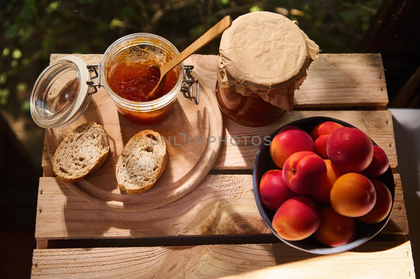 Overhead view of delicious ripe red apricots in a blue ceramic bowl on a wooden crate with jar of apricot confiture and slices of homemade freshly baked whole grain bread. Rustic style. Still life