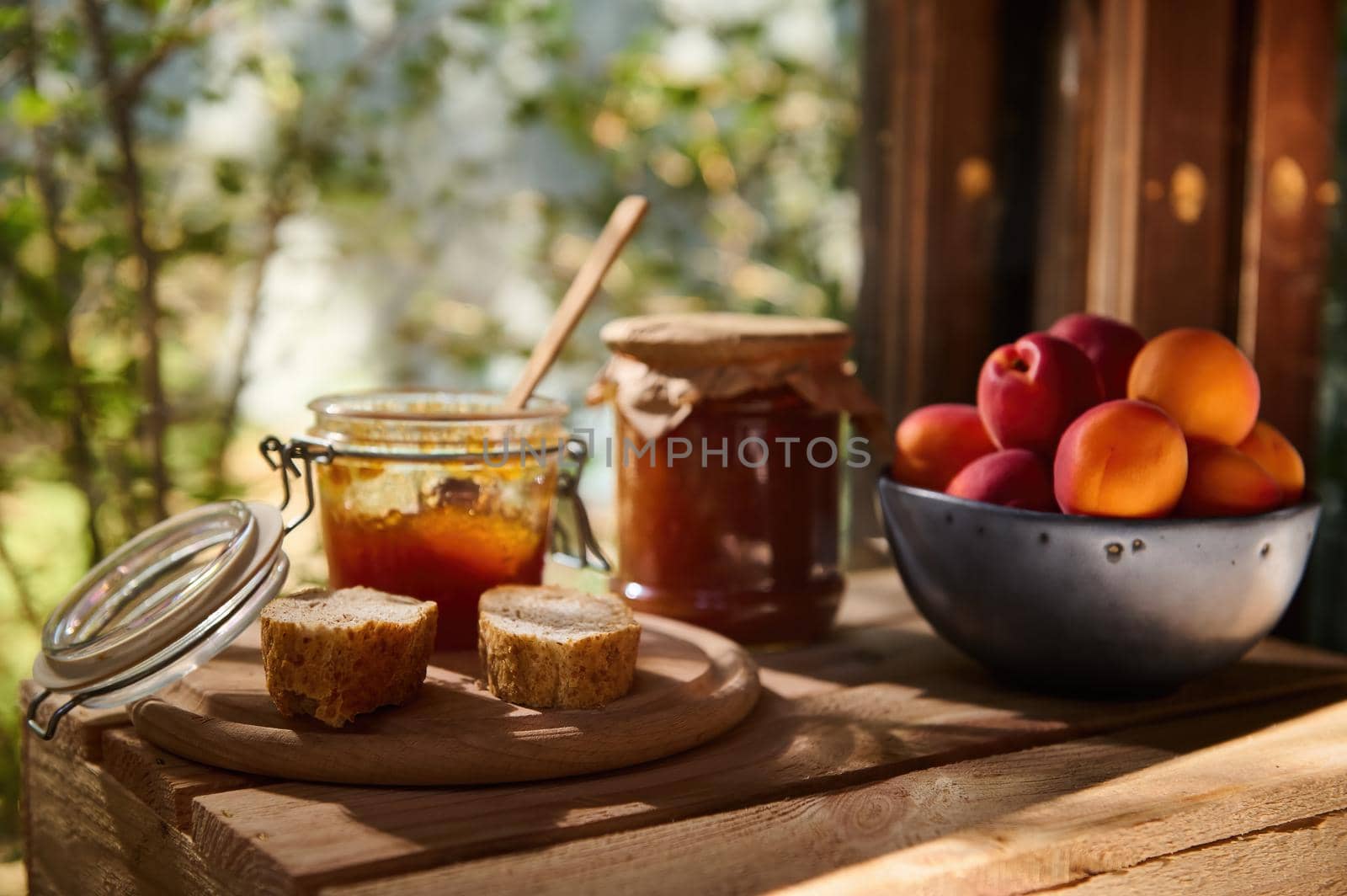 Food composition of delicious ripe apricots in blue ceramic bowl on wooden table with jar of apricot jam and slices of homemade bread, against the background of rural garden. Rustic style. Still life