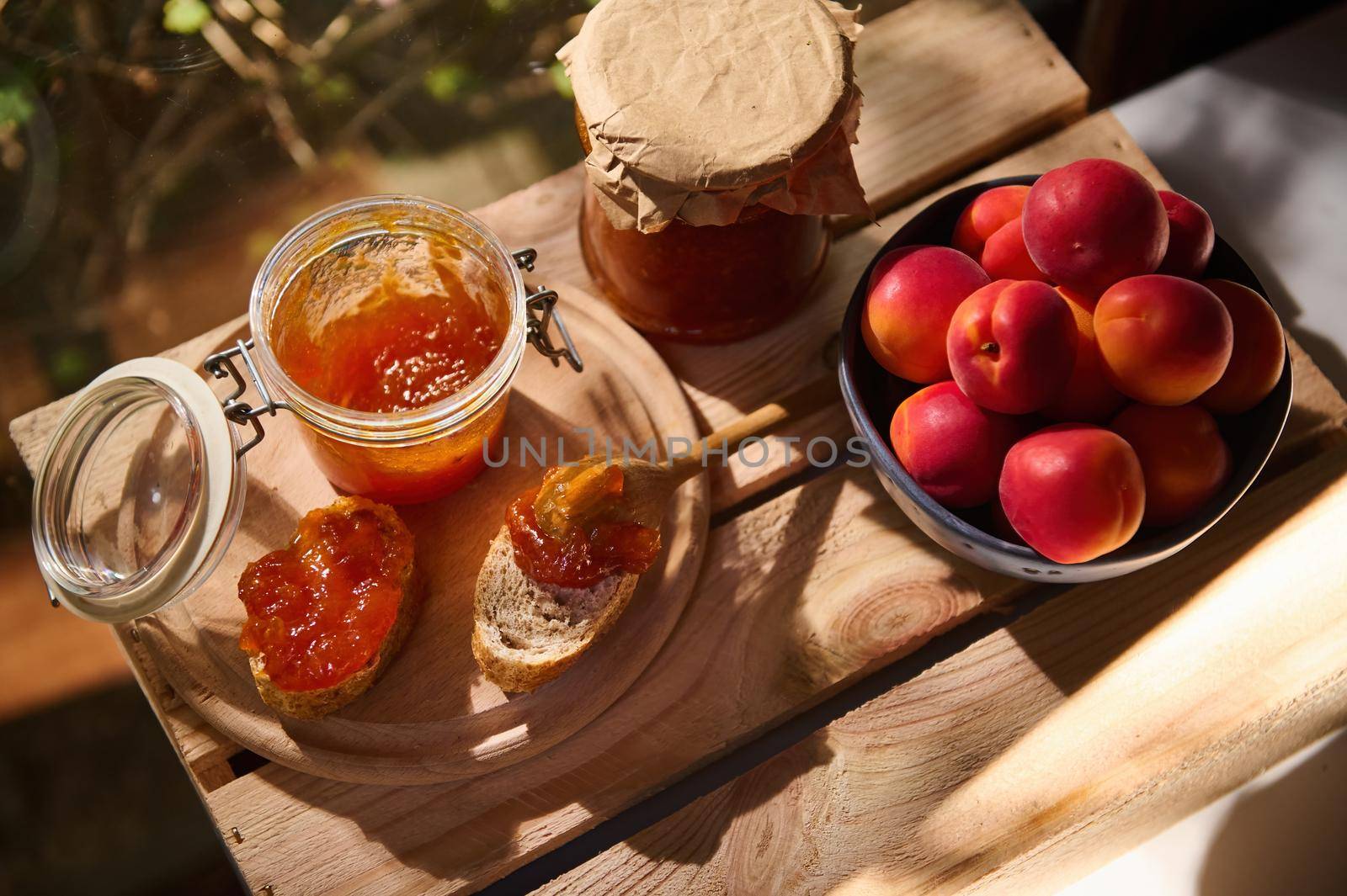 Top view of delicious ripe red apricots in a blue ceramic bowl on a wooden table with a glass jar of homemade apricot confiture and slices of freshly baked whole grain bread. Rustic style. Still life