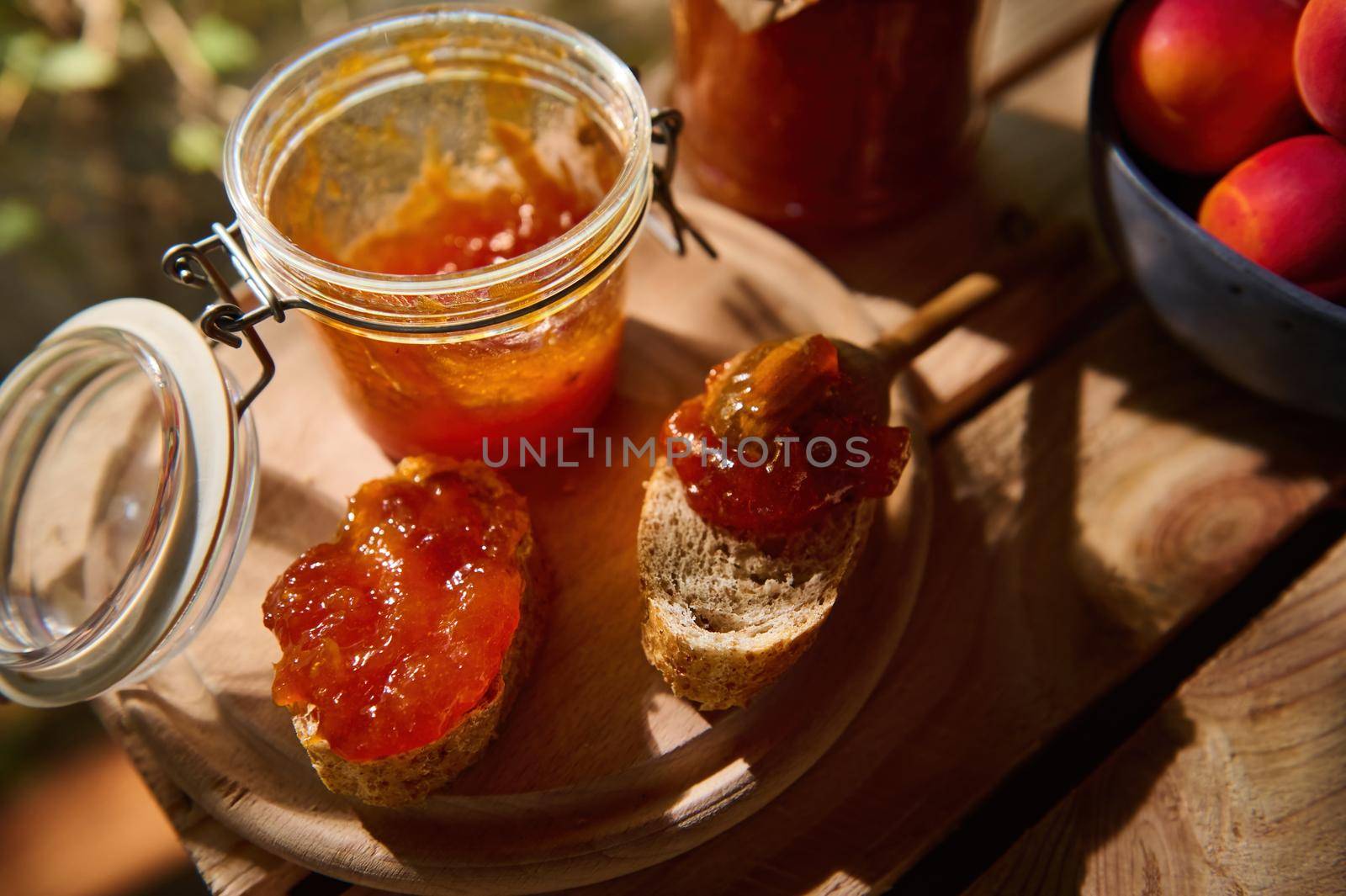 Top view of a homemade jam, slices of whole grain bread on a wooden board and partial view of ripe sweet apricots in a blue ceramic bowl, on a wooden crate. Canned food, sweet dessert. Still life
