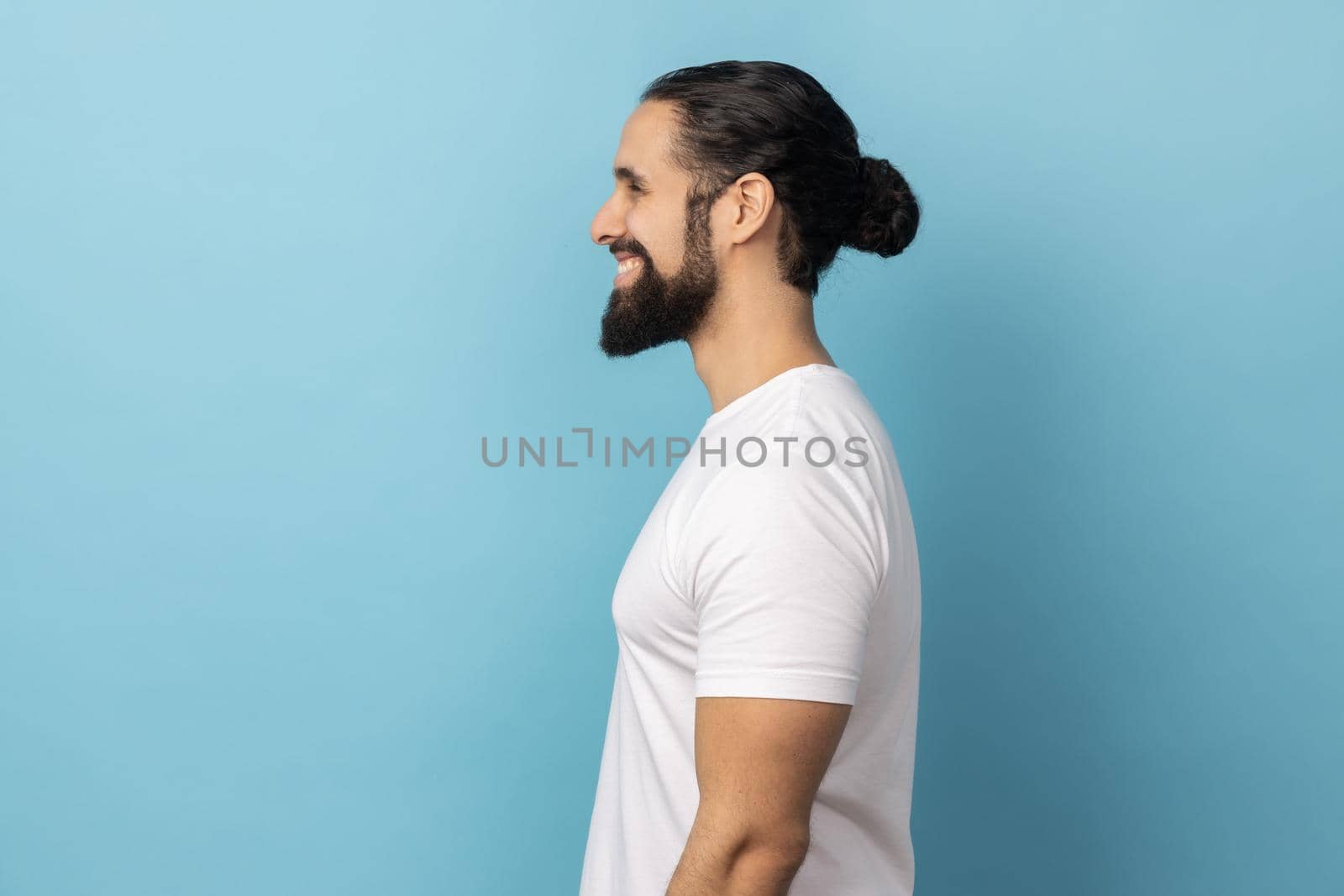 Side view of bearded handsome man wearing white T-shirt standing looking ahead with satisfied face and smiling, expressing happiness. Indoor studio shot isolated on blue background.