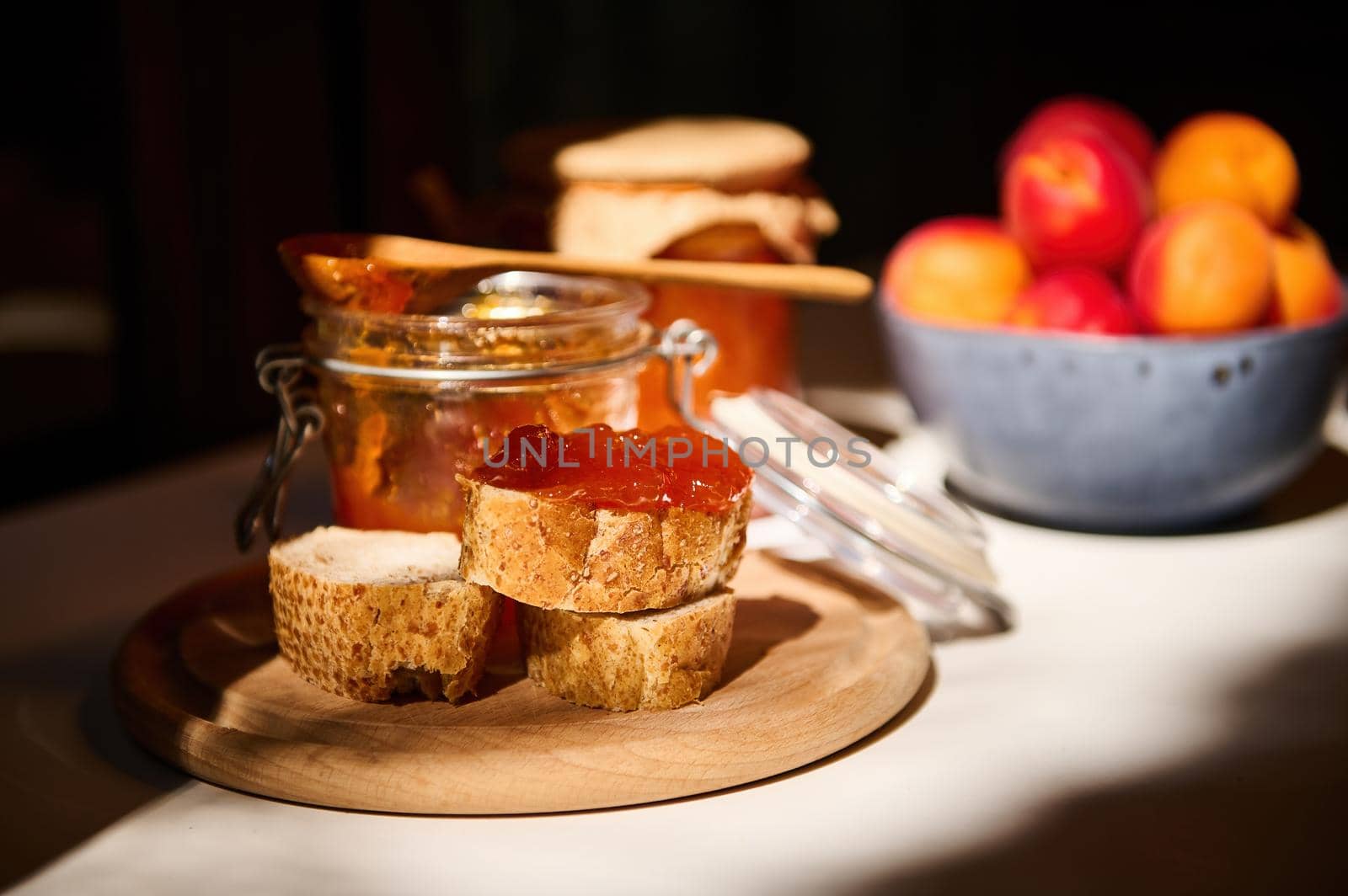 Daylight falls on part of the table. Sliced whole grain bread with jam on the background of closed jars of homemade apricot confiture and ripe sweet fruits in a blue ceramic bowl. Rustic still life