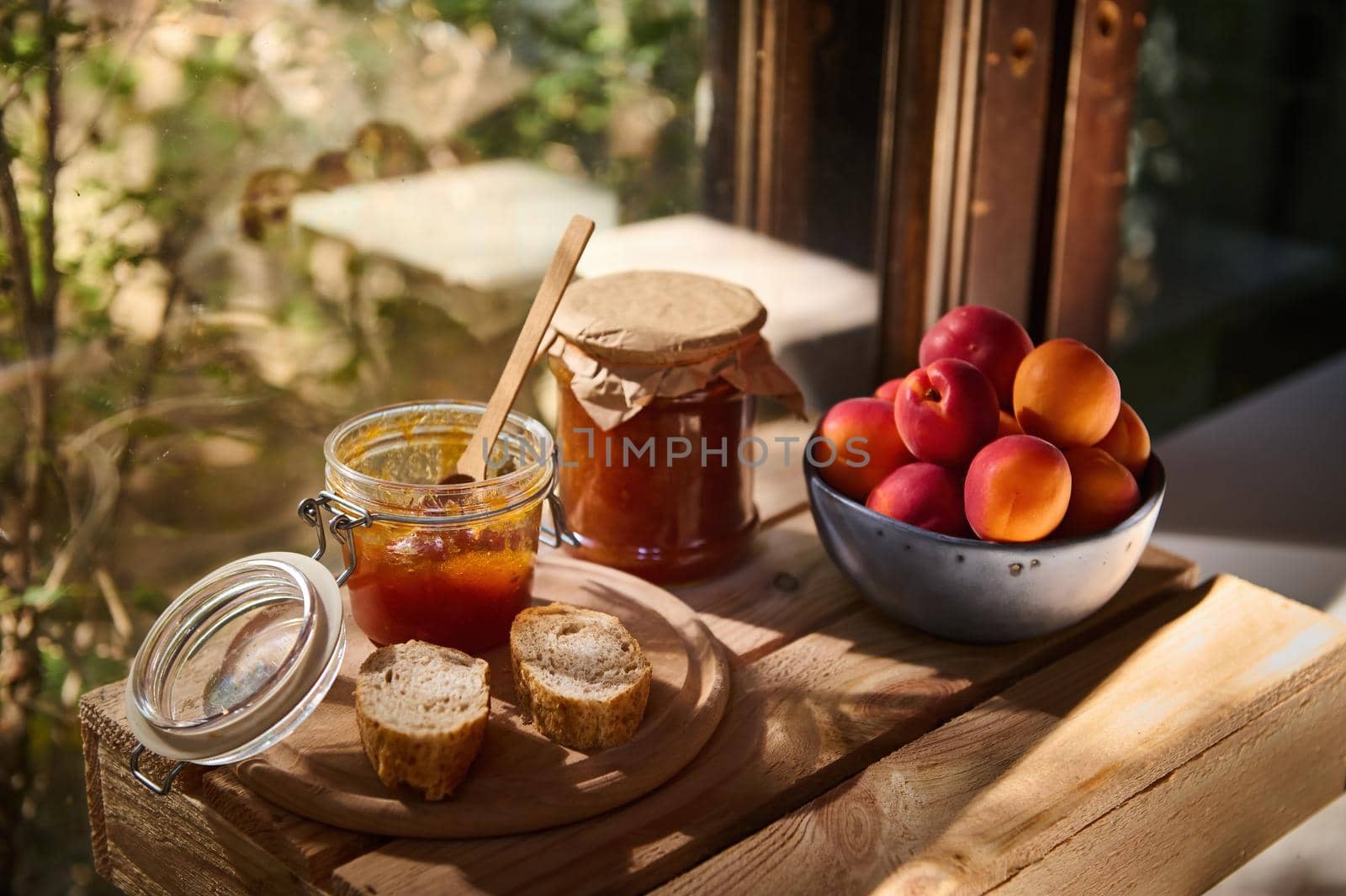 Still life. Canned products. Homemade jam and marmalade in a glass jar, slices of a freshly baked whole grain bread and sweet ripe organic apricots against a wooden window overlooking a rural garden