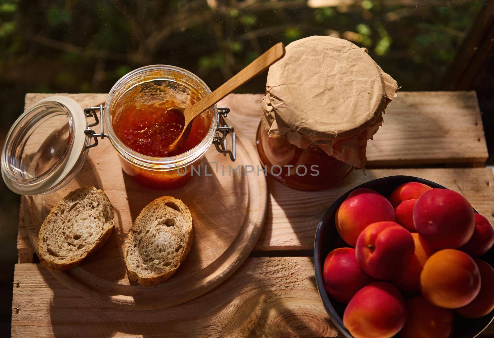 Top view. Still life. Homemade apricot jam in a glass jar with a wooden spoon, slices of whole grain bread on wooden crate near a blue ceramic bowl with freshly picked red ripe apricots. Country style