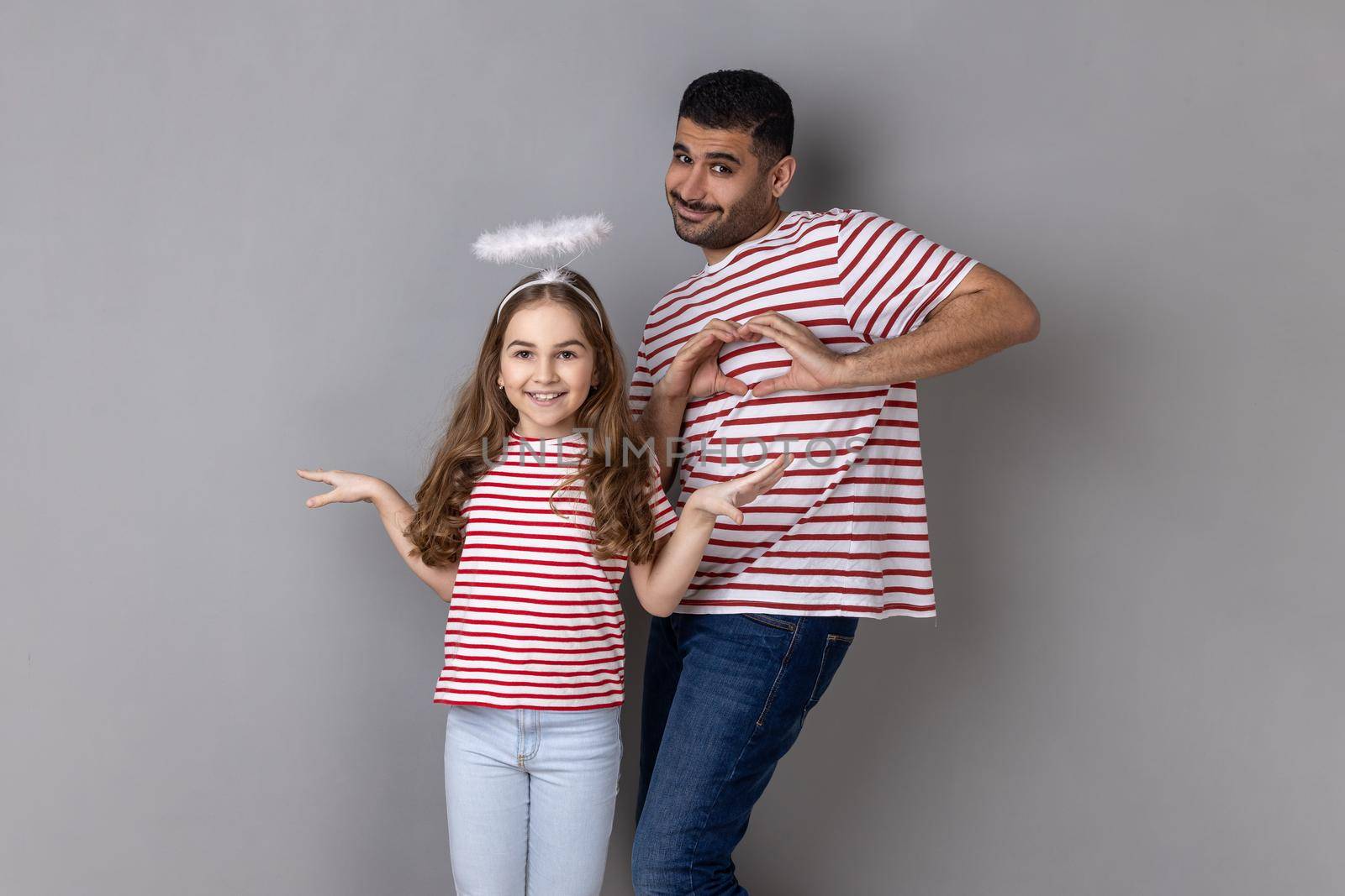 Portrait of optimistic father and daughter in striped T-shirts posing together, little girl with halo above head, man showing heart gesture with hands. Indoor studio shot isolated on gray background.