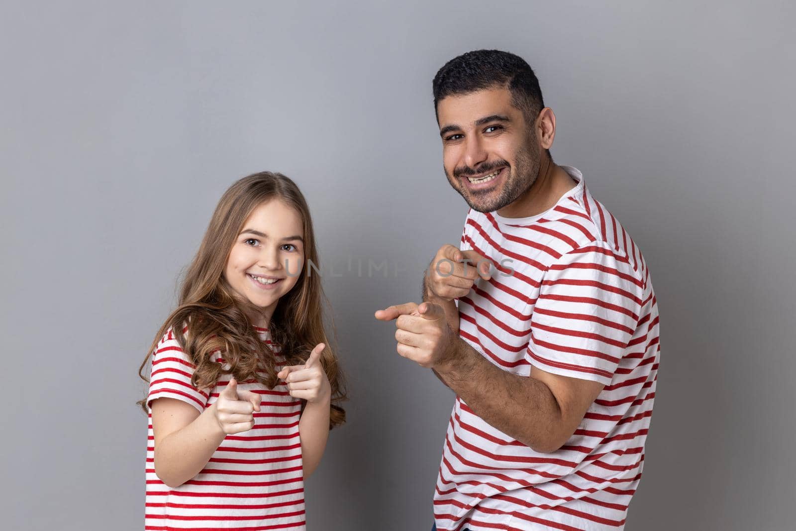 Portrait of optimistic happy father and daughter in striped T-shirts pointing fingers at camera, looking with charming smile, choosing you. Indoor studio shot isolated on gray background.