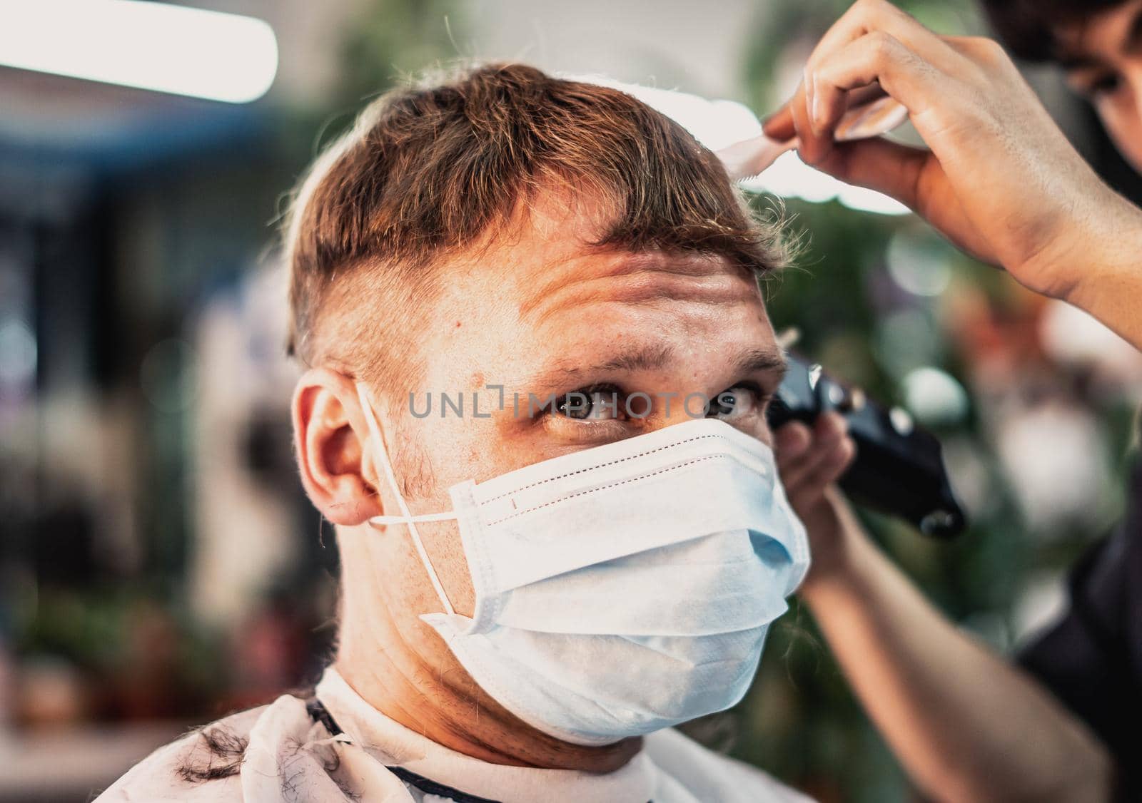 Lifestyle photo barbershop. Young man customer wears face mask, surprised dissatisfied look, wrinkled forehead. Hairdresser male hand work comb tool electric clipper. Short hair locks cut.