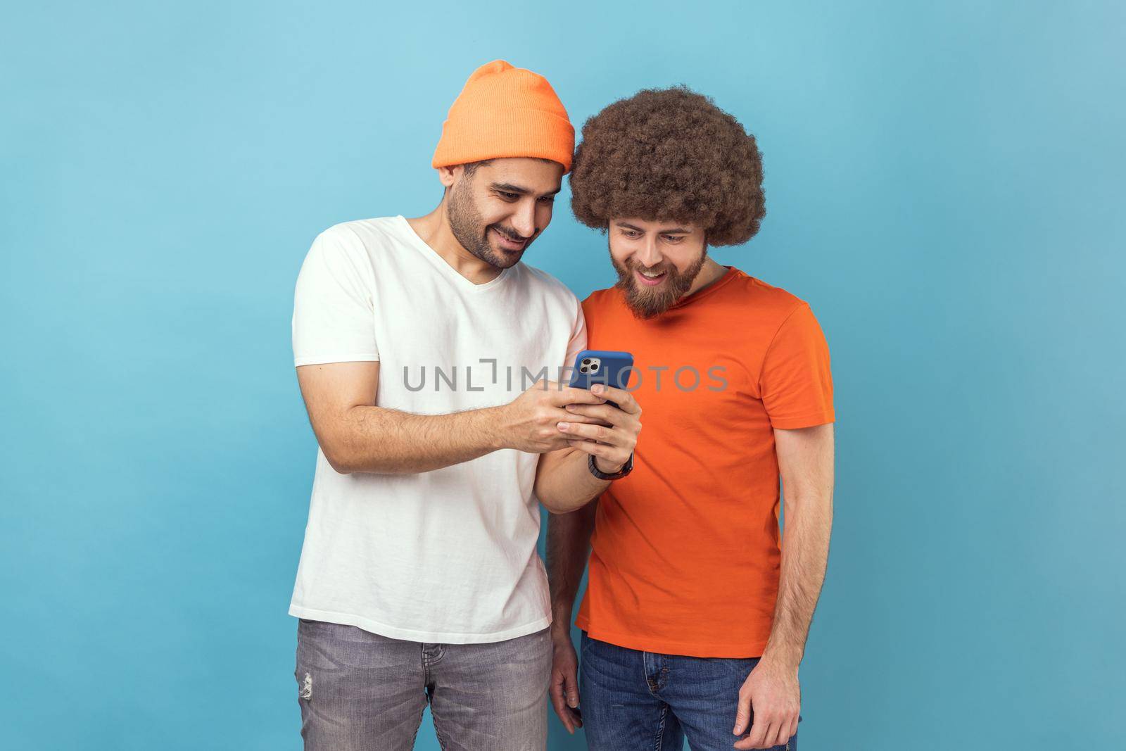 Portrait of two handsome optimistic young adult hipster men standing, using smartphone, reading positive news in social networks, smiling. Indoor studio shot isolated on blue background.
